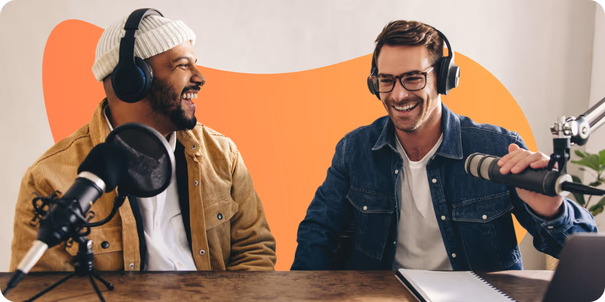 Two men wearing headphones laughing and speaking into microphones while recording a podcast.