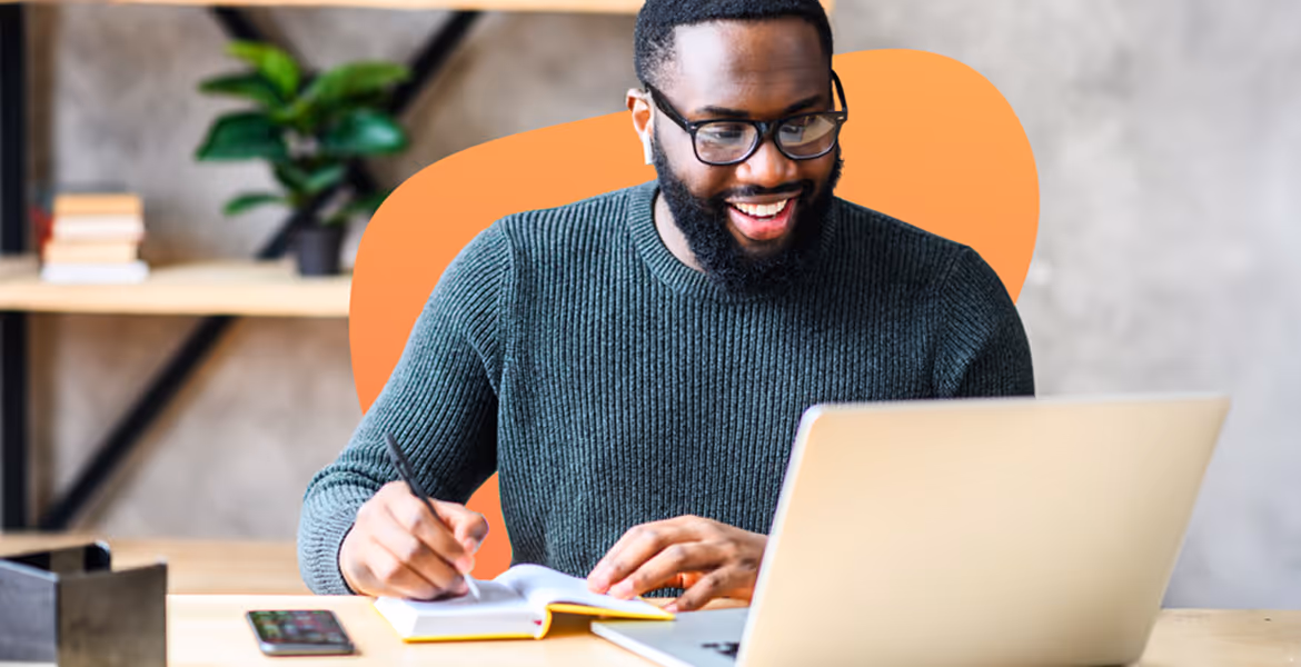 Smiling man wearing glasses and a green sweater writing in a notebook while looking at a laptop.
