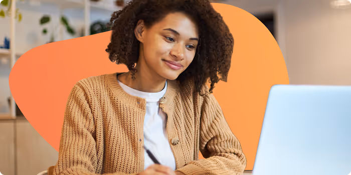 Young woman with curly hair wearing a brown cardigan, working and writing while looking at a laptop screen.