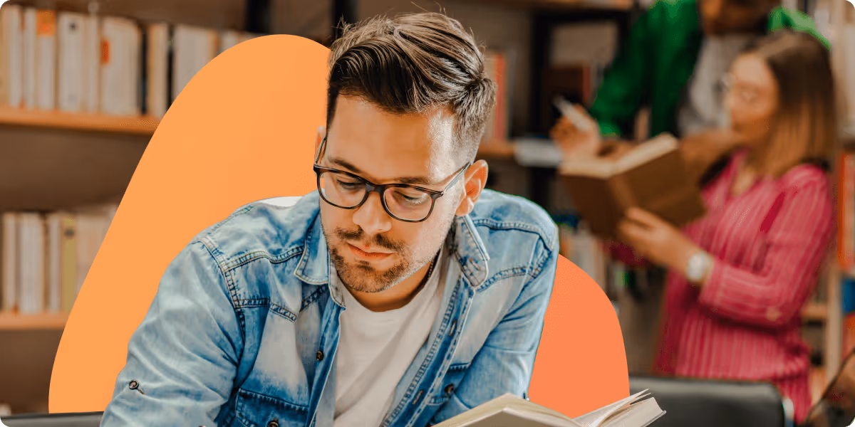Young man wearing glasses and a denim jacket reading a book in a library with a woman and man browsing books in the background.