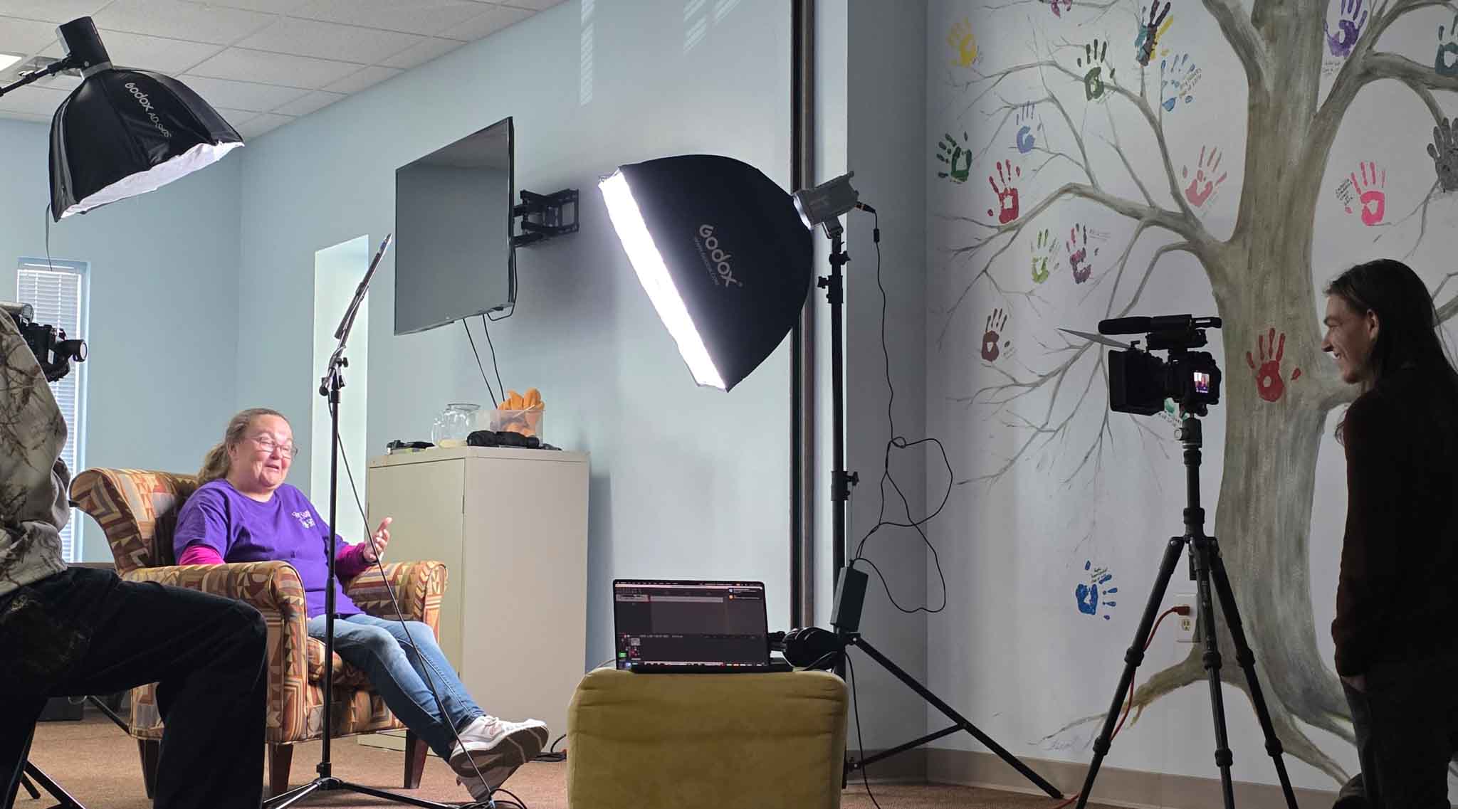 Video production setup for an interview with lighting, camera, and audio equipment in a community space.