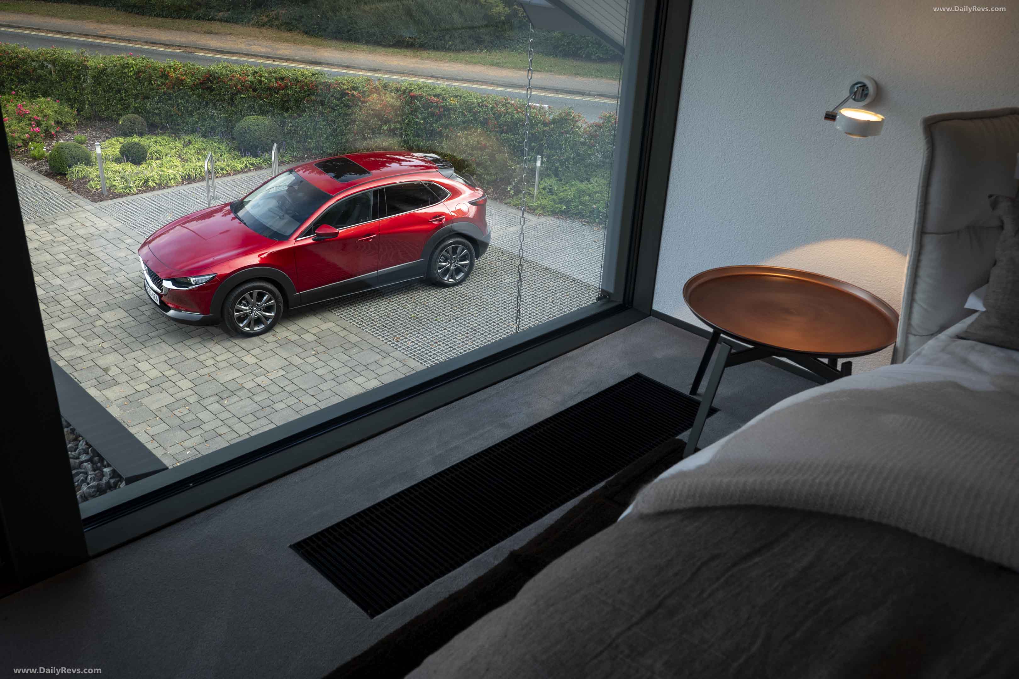 View from a modern bedroom with gray bedding and a round side table looking out a large window at a red crossover SUV parked on a stone driveway.