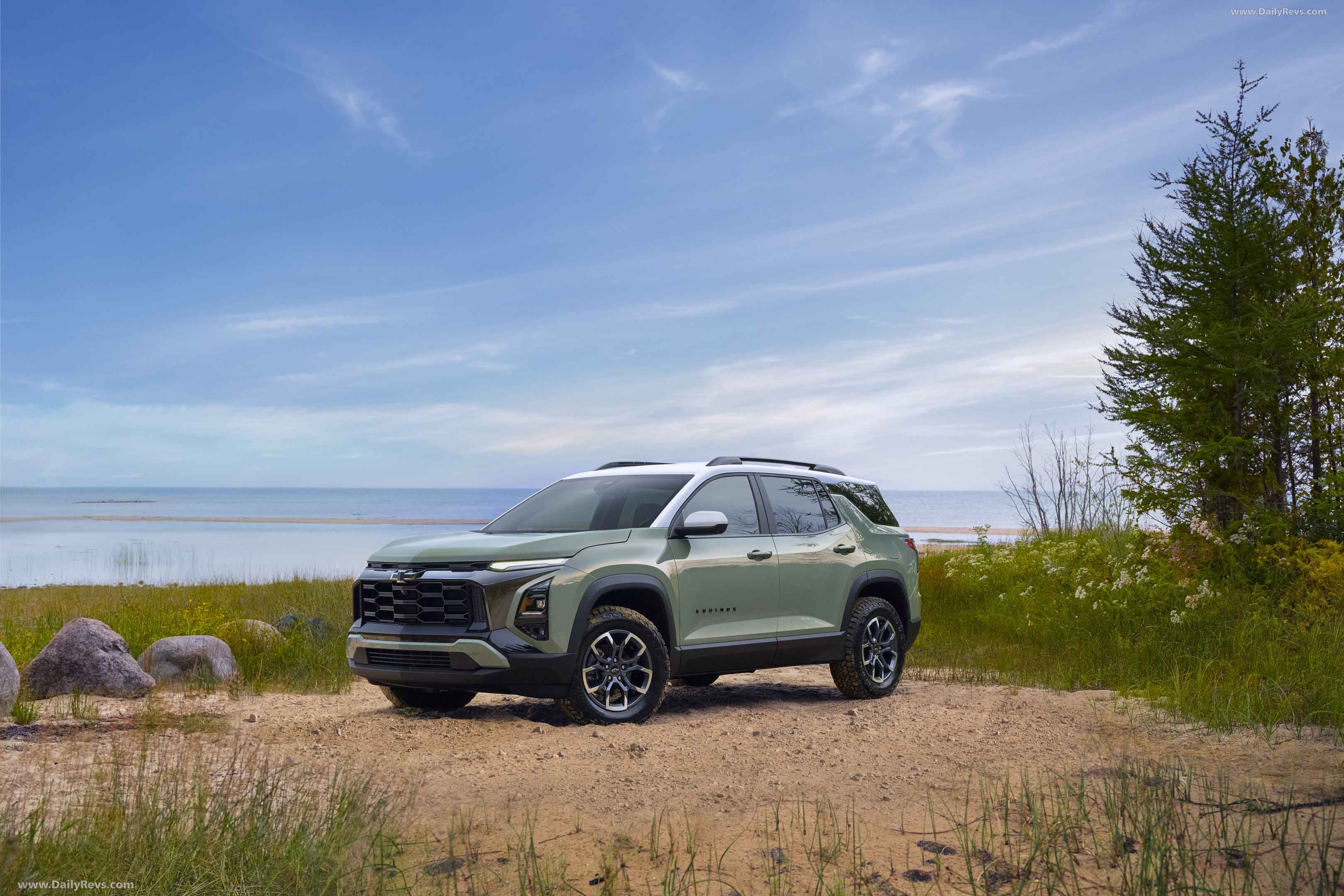 Green Chevrolet Equinox SUV parked on sandy ground near grassy bushes and a calm body of water under a blue sky.