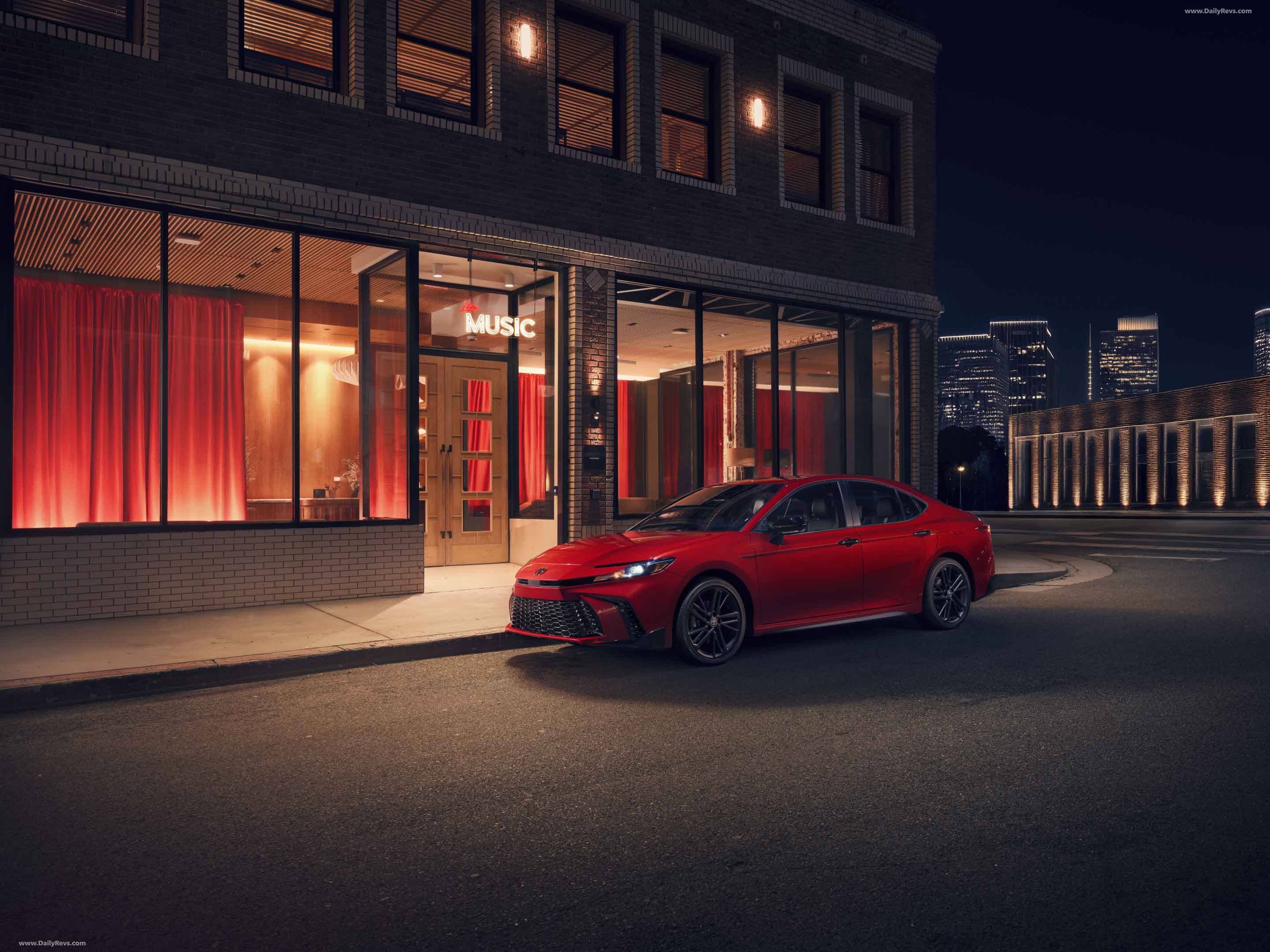 Red sedan parked on a city street at night in front of a building with large windows and red curtains, under a sign that reads 'MUSIC'.