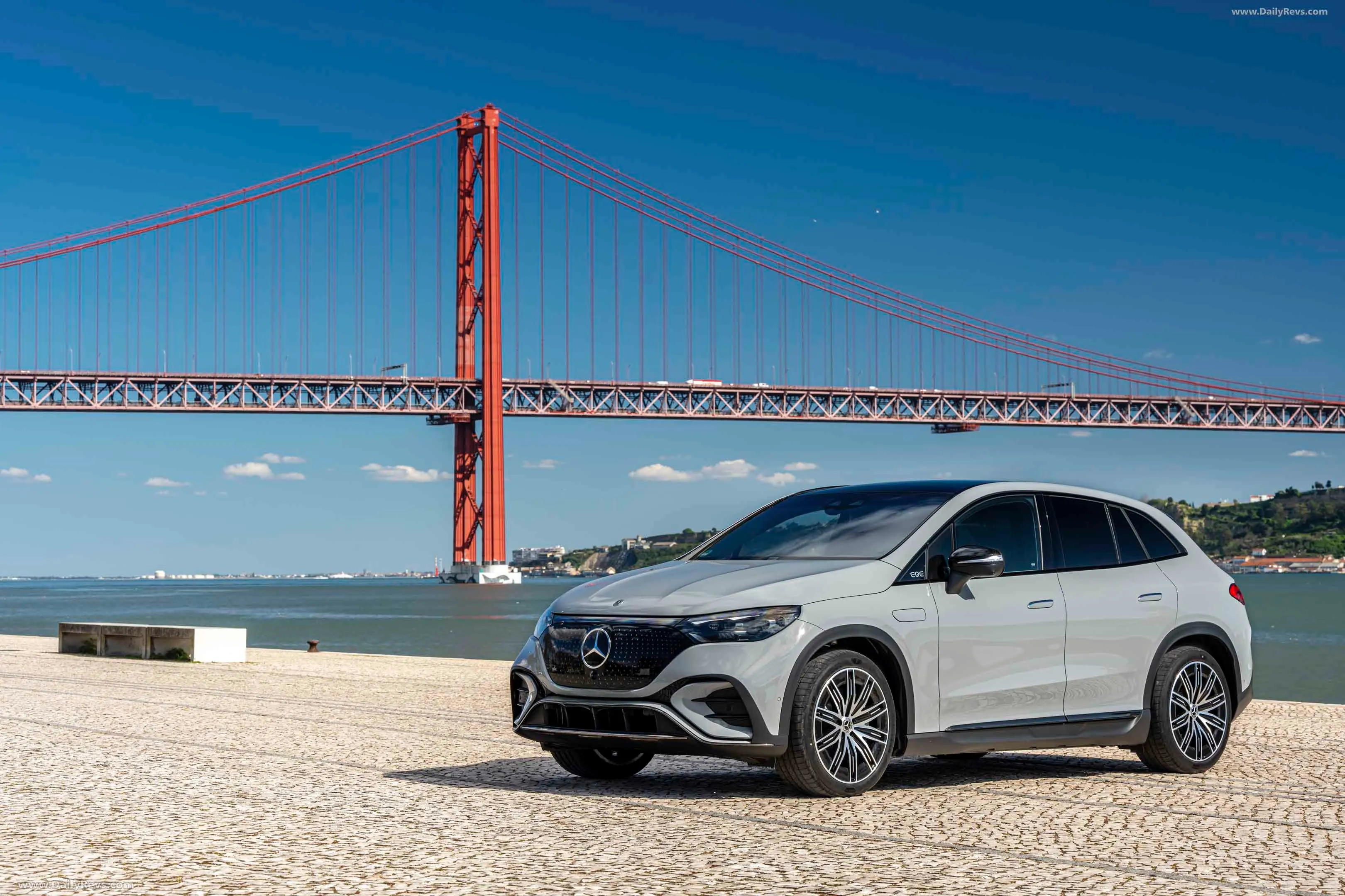 White Mercedes-Benz EQE SUV parked on a paved area with a red suspension bridge and water in the background under a clear blue sky.