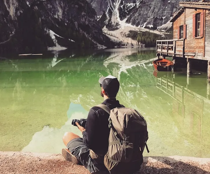 A person sitting by a crystal-clear lake, surrounded by mountains and a wooden cabin.