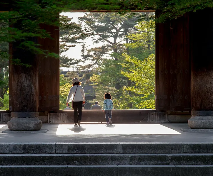 A mother and child walking through a temple gate with green trees in the background.
