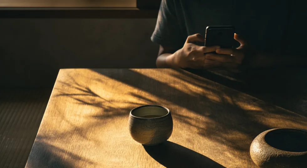 Person using a phone at a wooden table with soft sunlight.