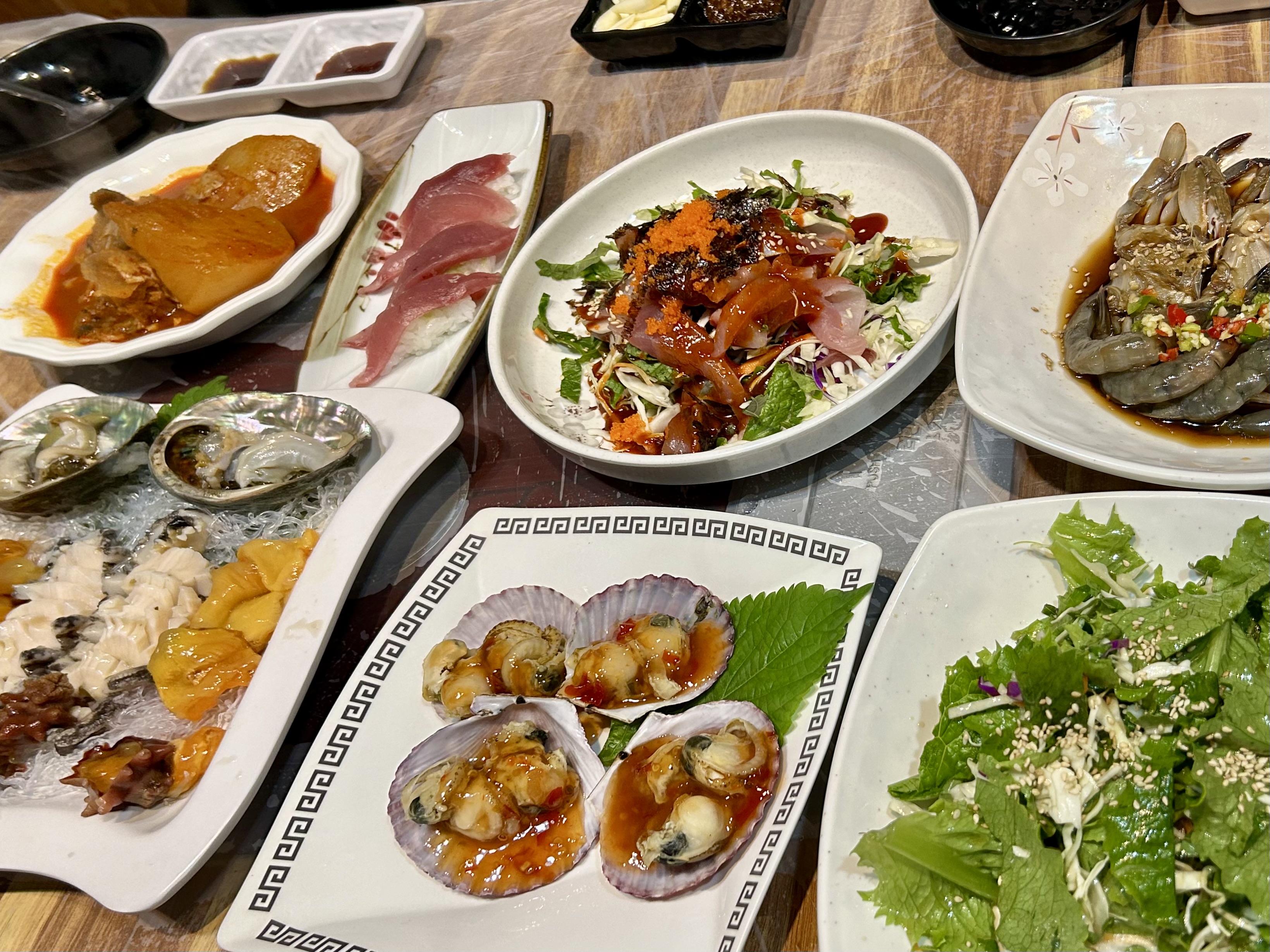 A Korean table set with assorted seafood dishes, including raw shellfish, scallops, marinated shrimp, salads, and small side plates.