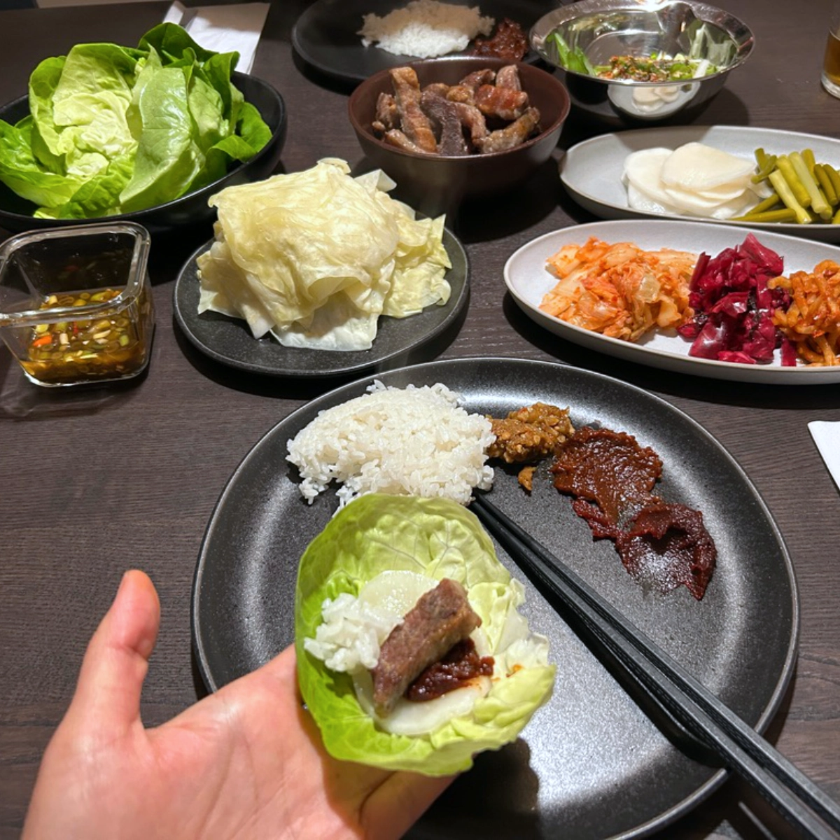 A hand holding a lettuce wrap filled with pork, rice, and sauce, with side dishes, kimchi, and condiments spread across a Korean barbecue table.