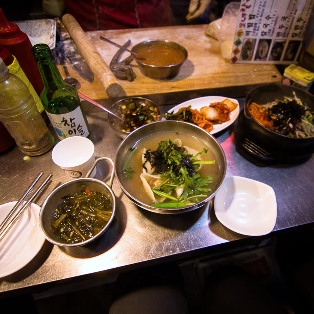 A metal market counter with a bowl of manduguk topped with seaweed and greens, side dishes of kimchi and greens, a bottle of soju, and a stone pot of bibimbap in the background.