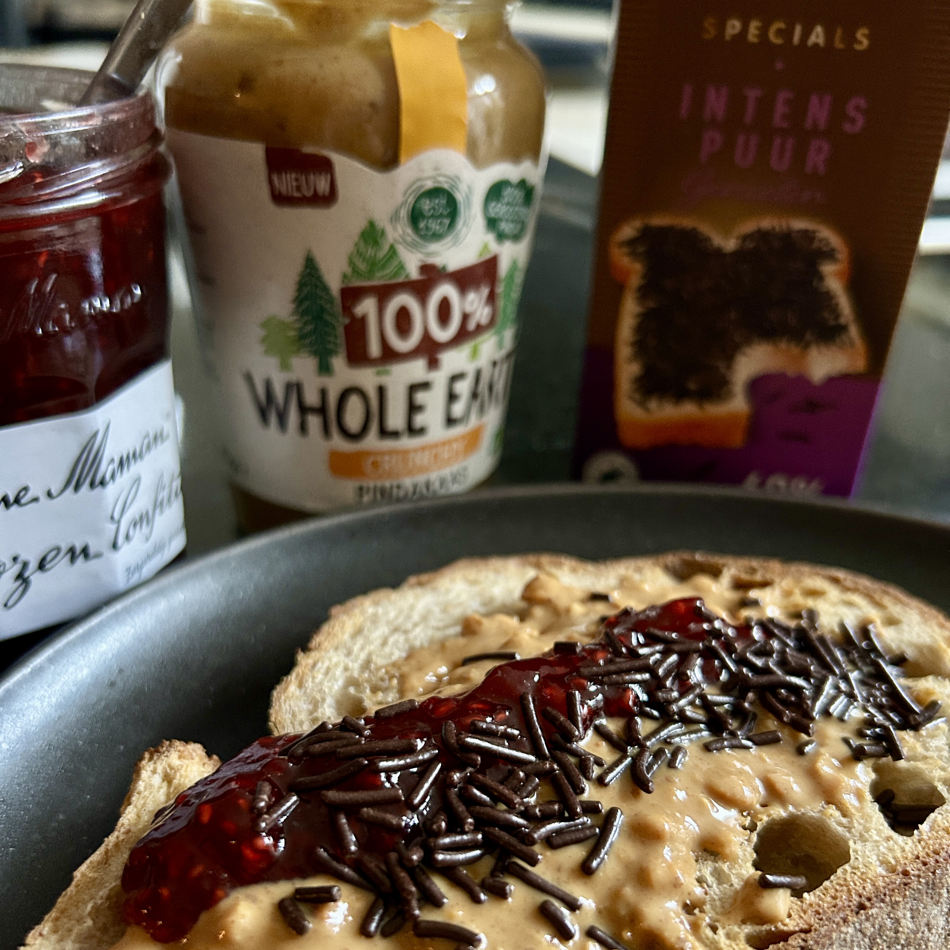 Slice of bread spread with peanut butter and strawberry jam, topped with chocolate sprinkles (hagelslag), with jars of jam and peanut butter in the background.