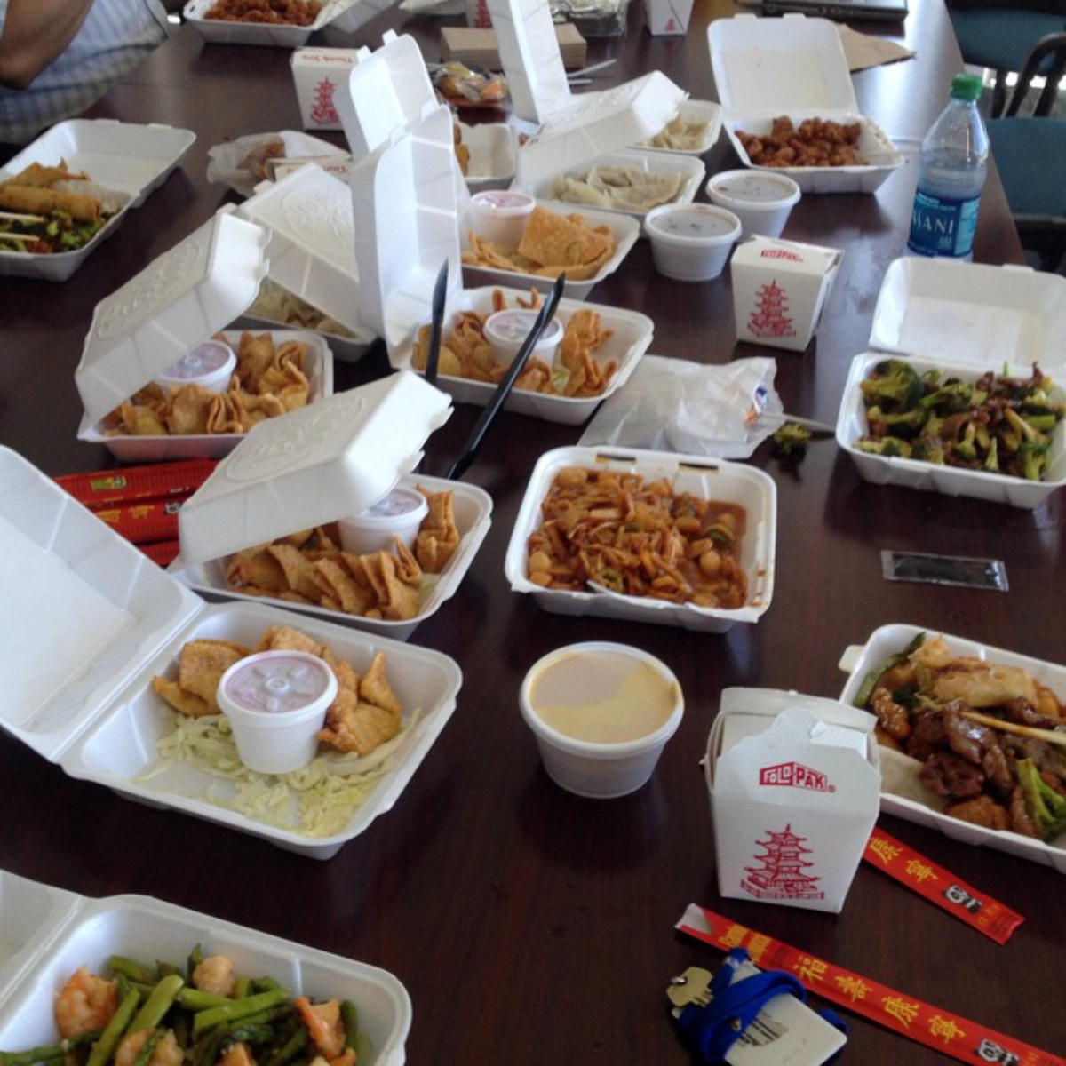 White Chinese American takeout containers filled with dishes such as fried rice, lo mein, broccoli with shrimp, dumplings, and sweet-and-sour items spread across a large office table with chopsticks and sauce cups visible.