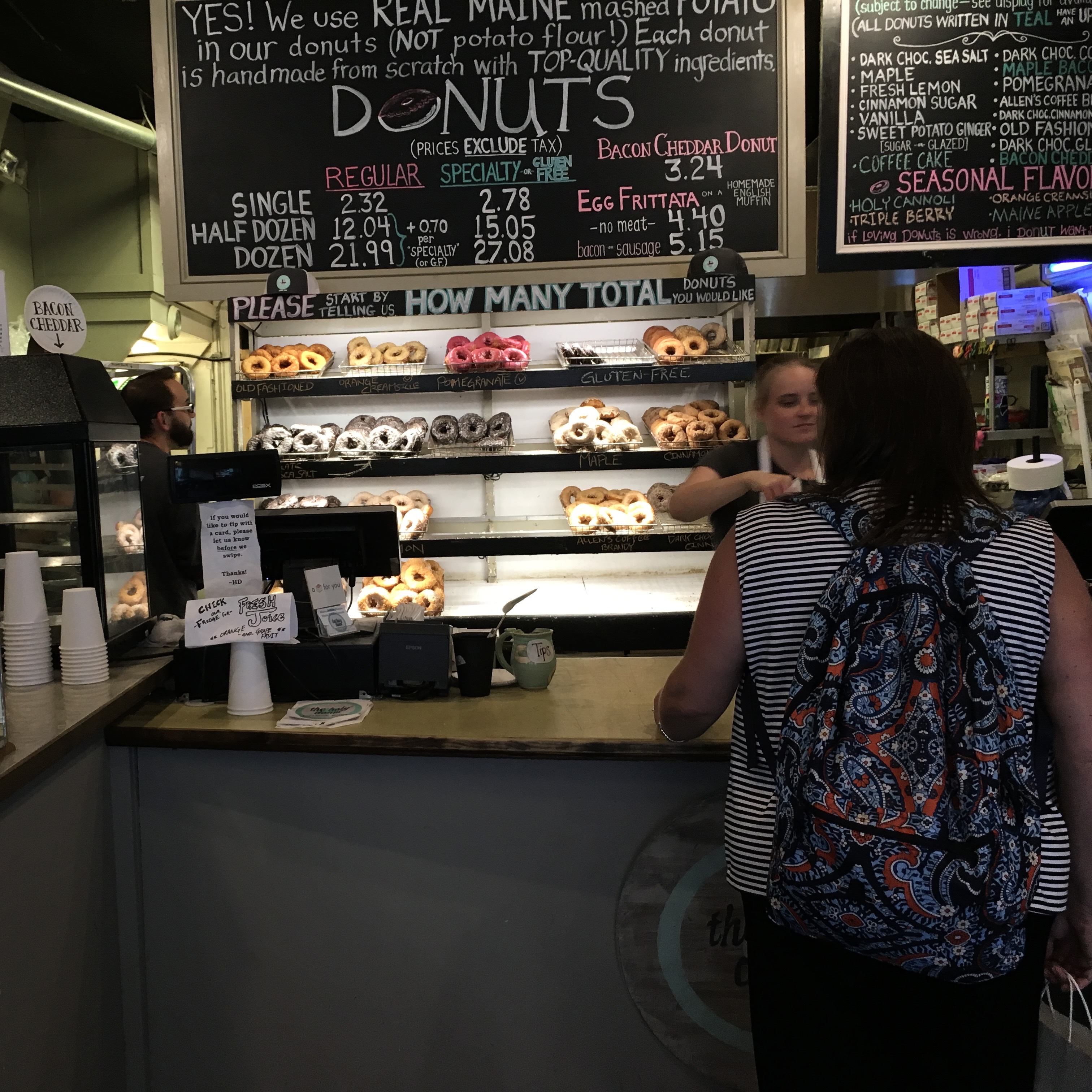 Interior of an American donut shop with a chalkboard menu listing single, half dozen, and dozen prices above shelves of donuts, highlighting the boxed quantities meant for sharing.