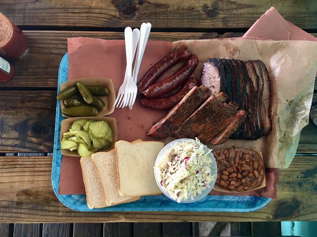 A Texas barbecue tray with sliced brisket, sausage, pickles, coleslaw, beans, and white bread.