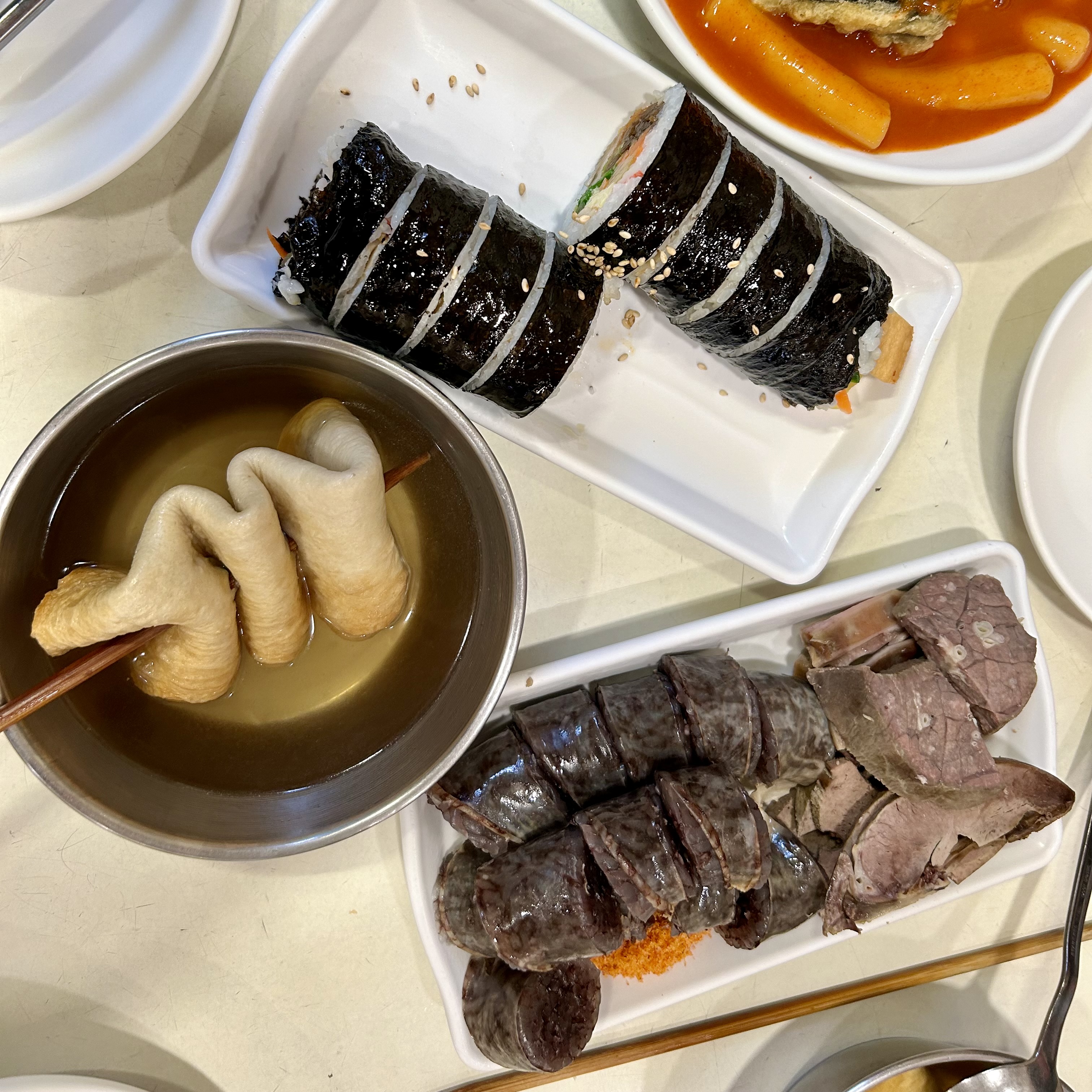 Korean blood sausage (sundae) with liver and assorted cuts, served with fish cake soup, gimbap, and tteokbokki on a table.