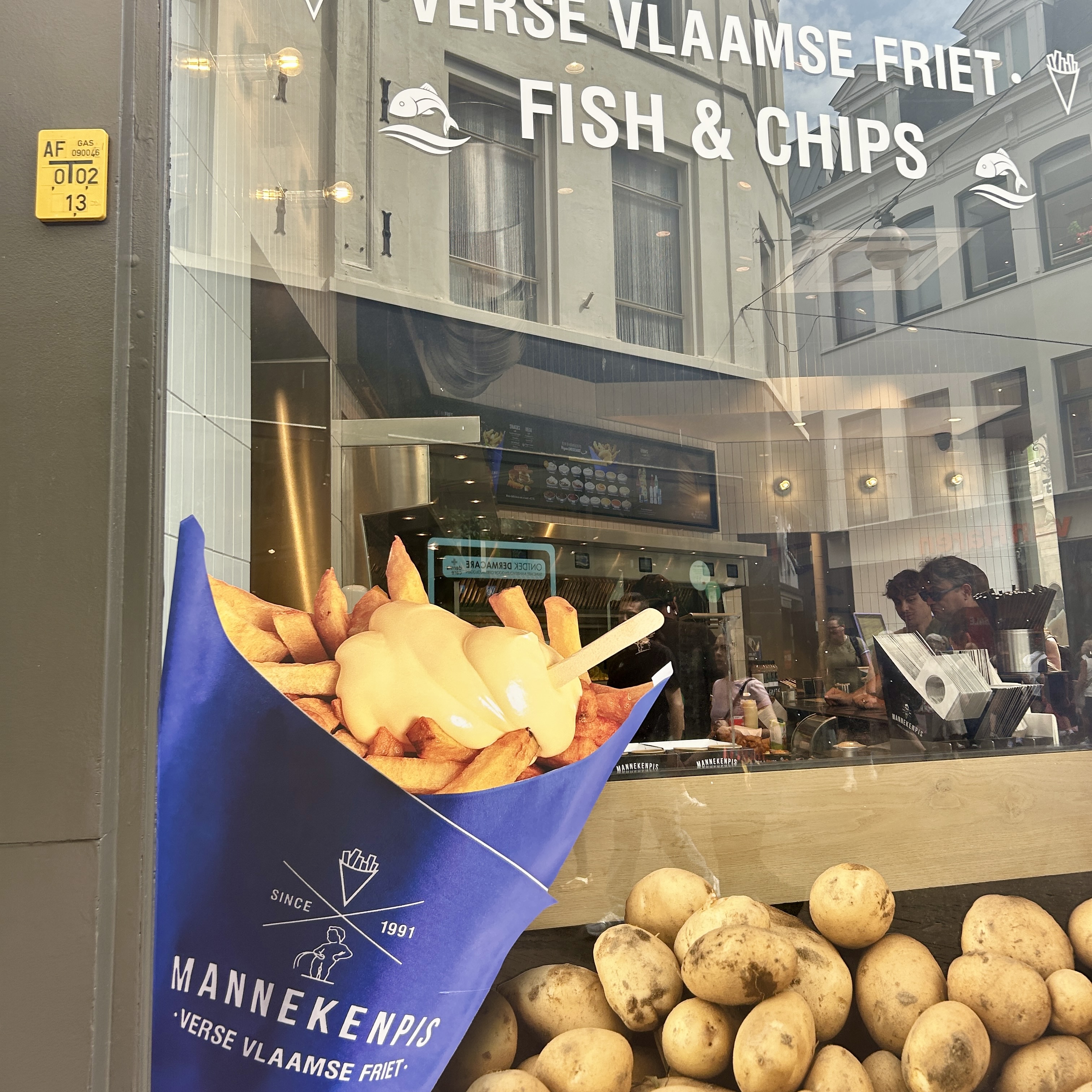 Thick-cut Flemish fries in a paper cone topped with mayonnaise, with raw potatoes displayed below in a shop window.