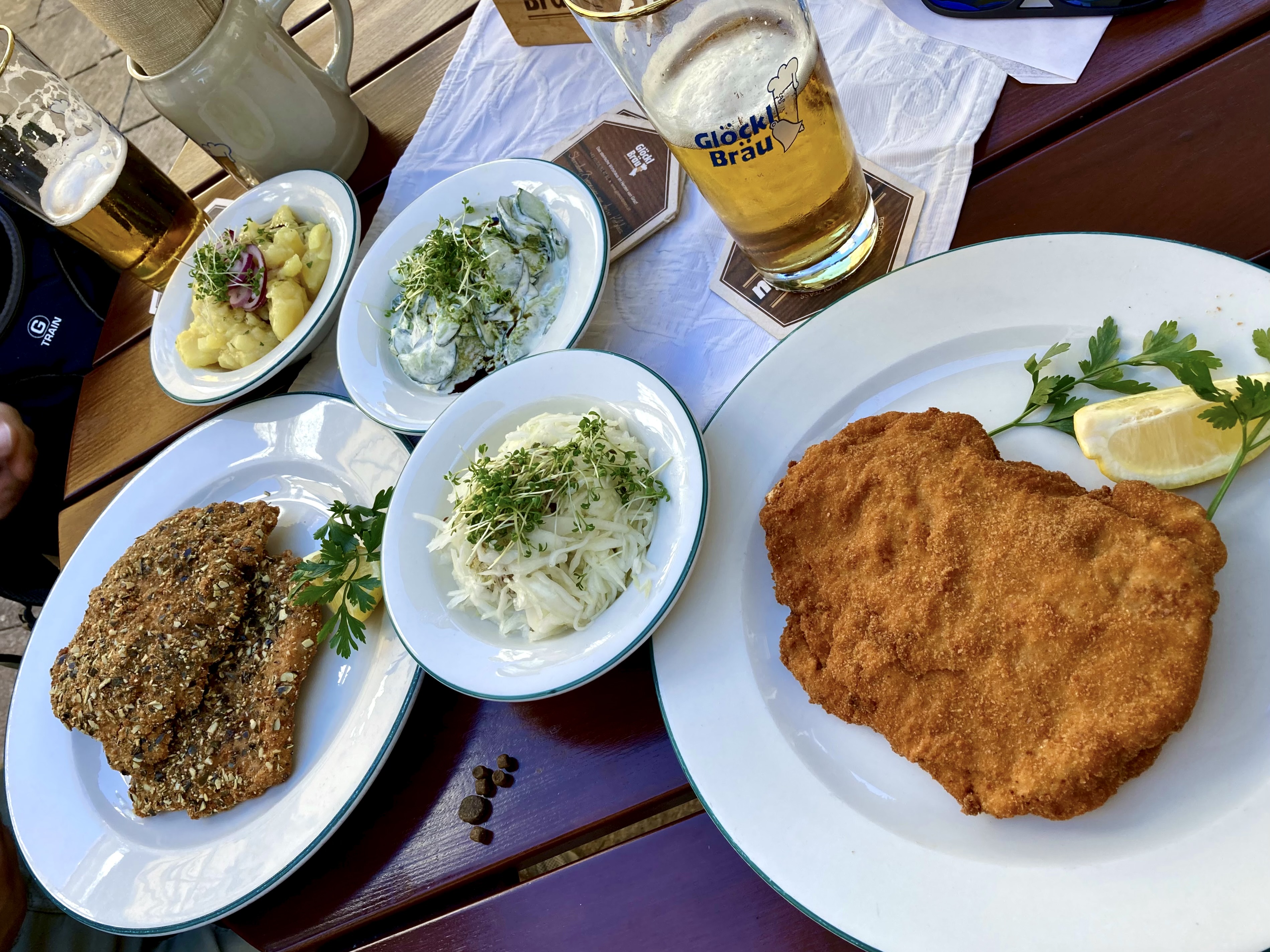 Breaded veal schnitzel with lemon, served alongside small side dishes and beer on an outdoor table.