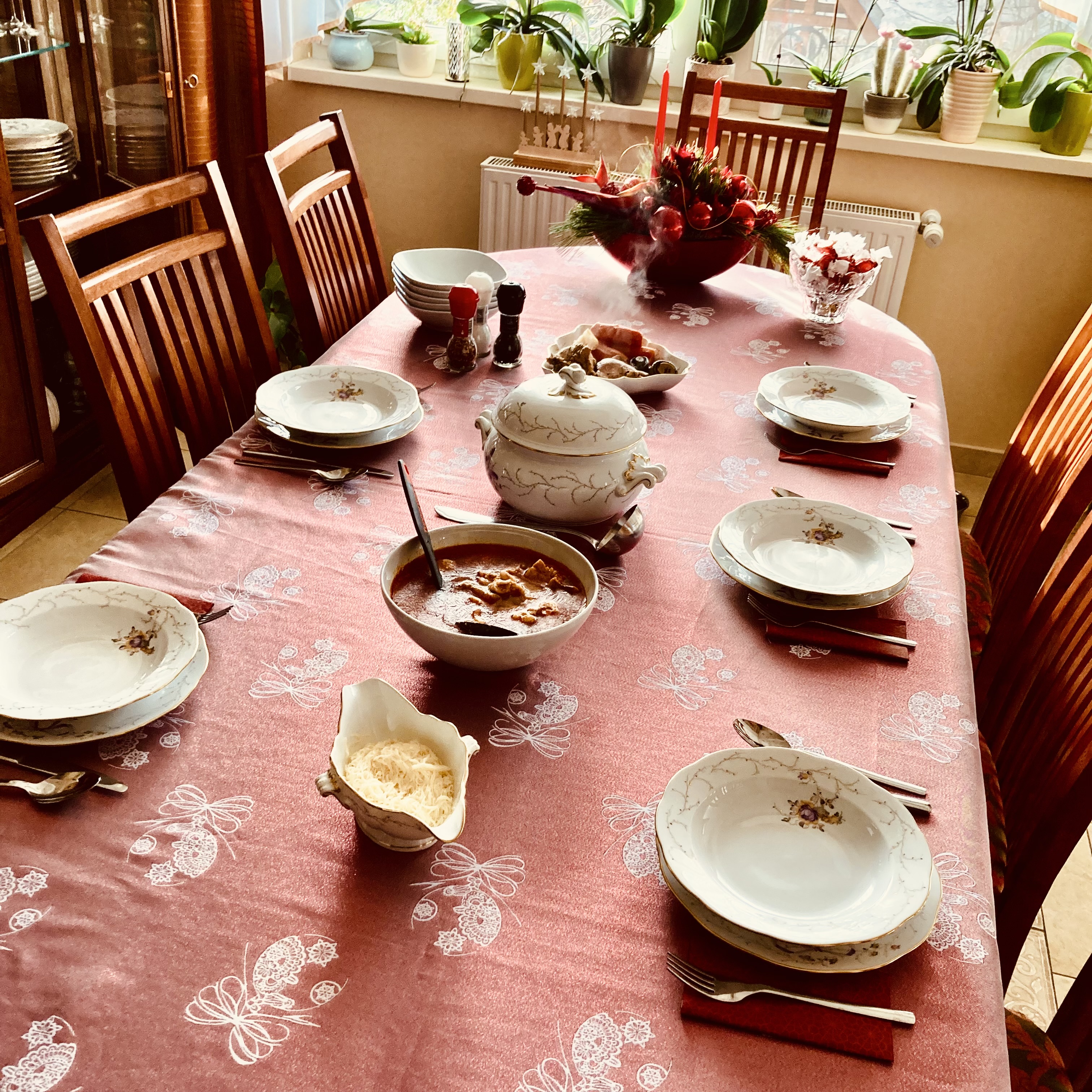 A dining table set for Christmas, with a red-orange soup in a serving bowl, a white porcelain tureen, and floral-patterned china. A red floral centerpiece and candles sit near the window.