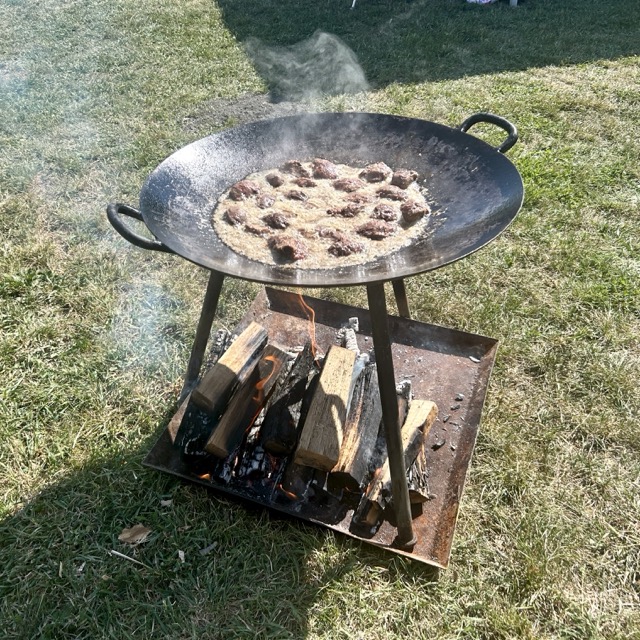 Chunks of fish frying in oil on a large iron pan over an open wood fire outdoors, with smoke rising above.