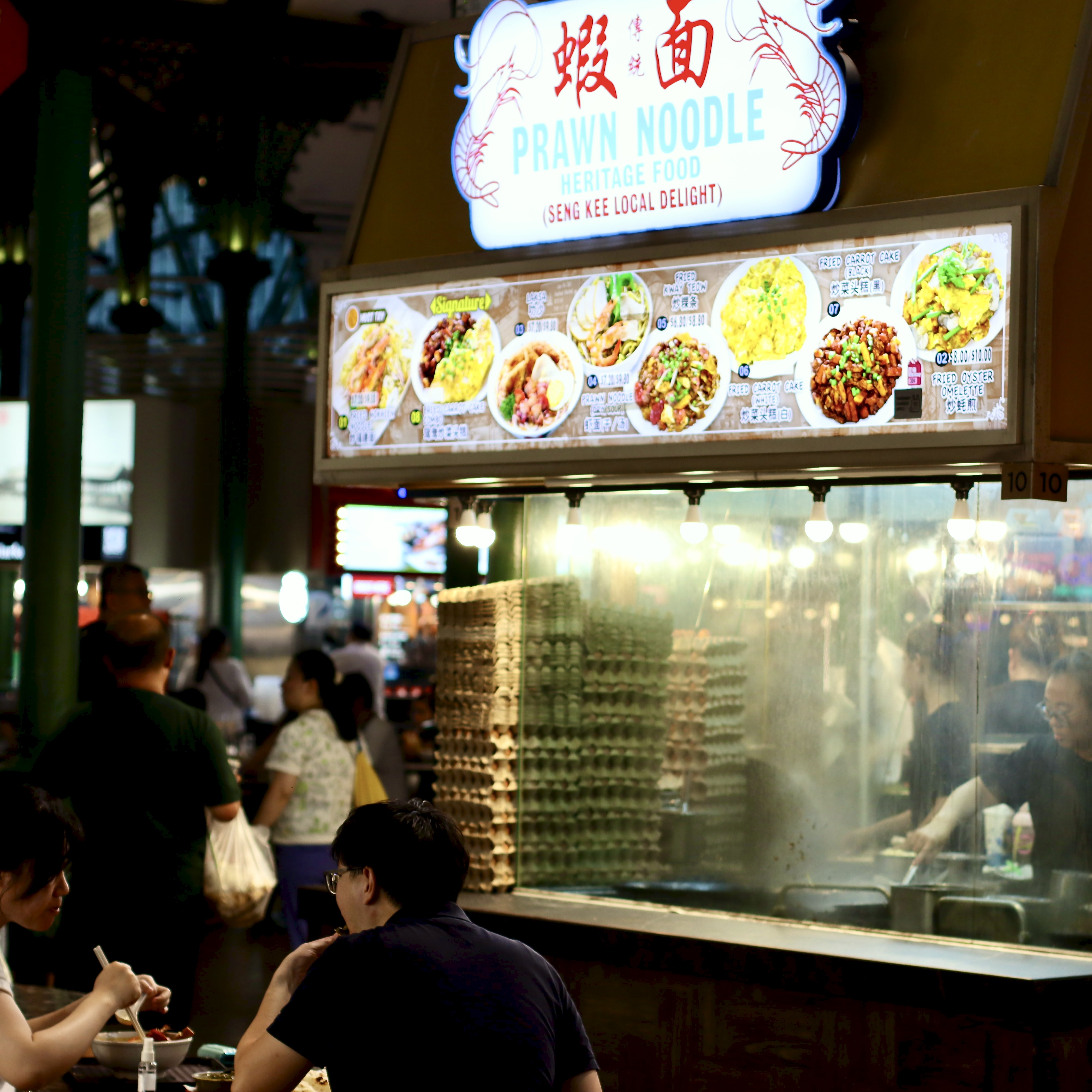 A lit hawker stall sign reading "Prawn Noodle Heritage Food" above a steaming open kitchen, with stacked egg trays and a cook working behind the counter.