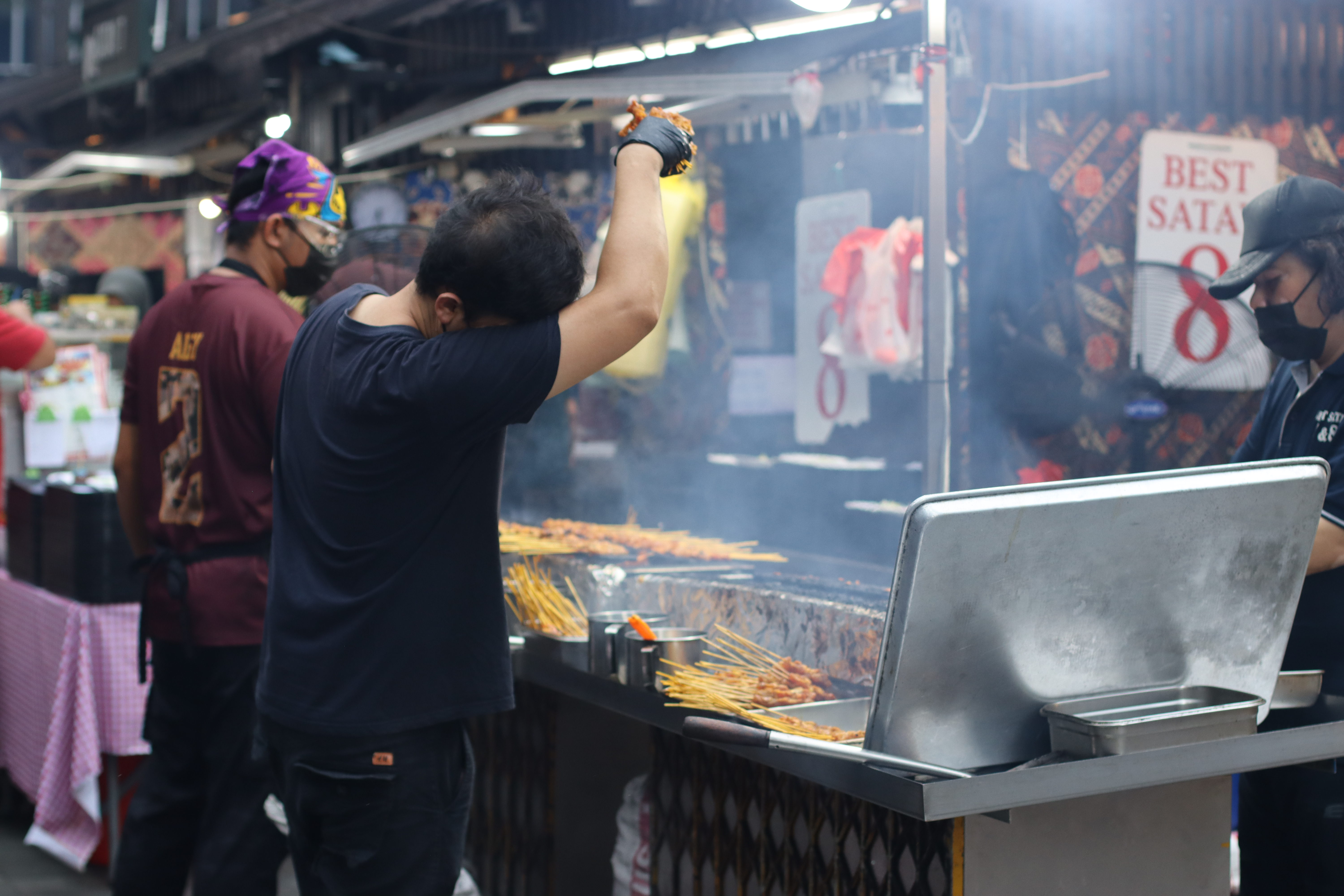 A cook turning satay skewers on a large outdoor charcoal grill, with smoke rising and rows of skewers lined up across the grate. A "Best Satay" sign visible in the background.