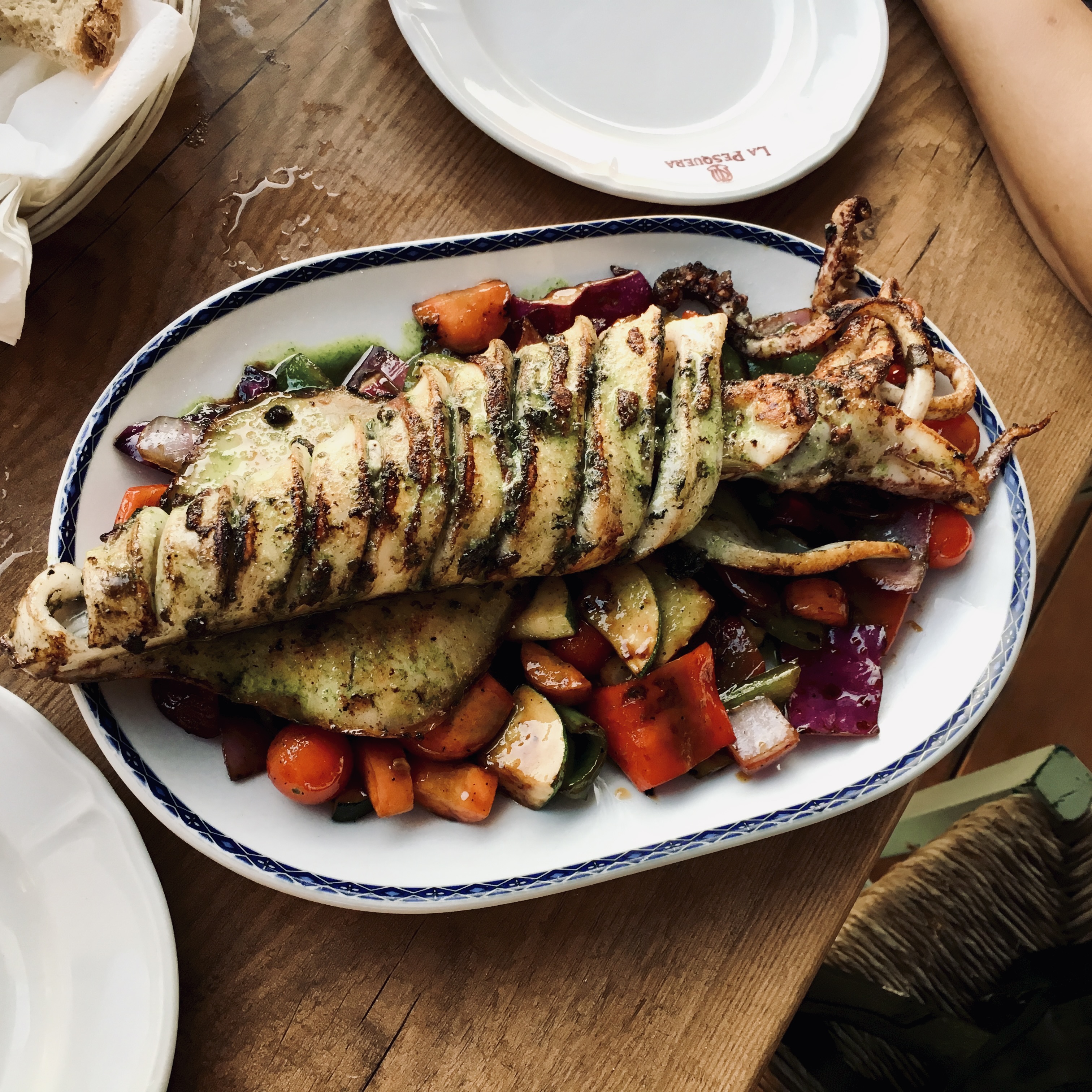 A whole grilled squid, scored and charred, drizzled with green herb sauce and served on roasted vegetables in a blue-rimmed oval plate.