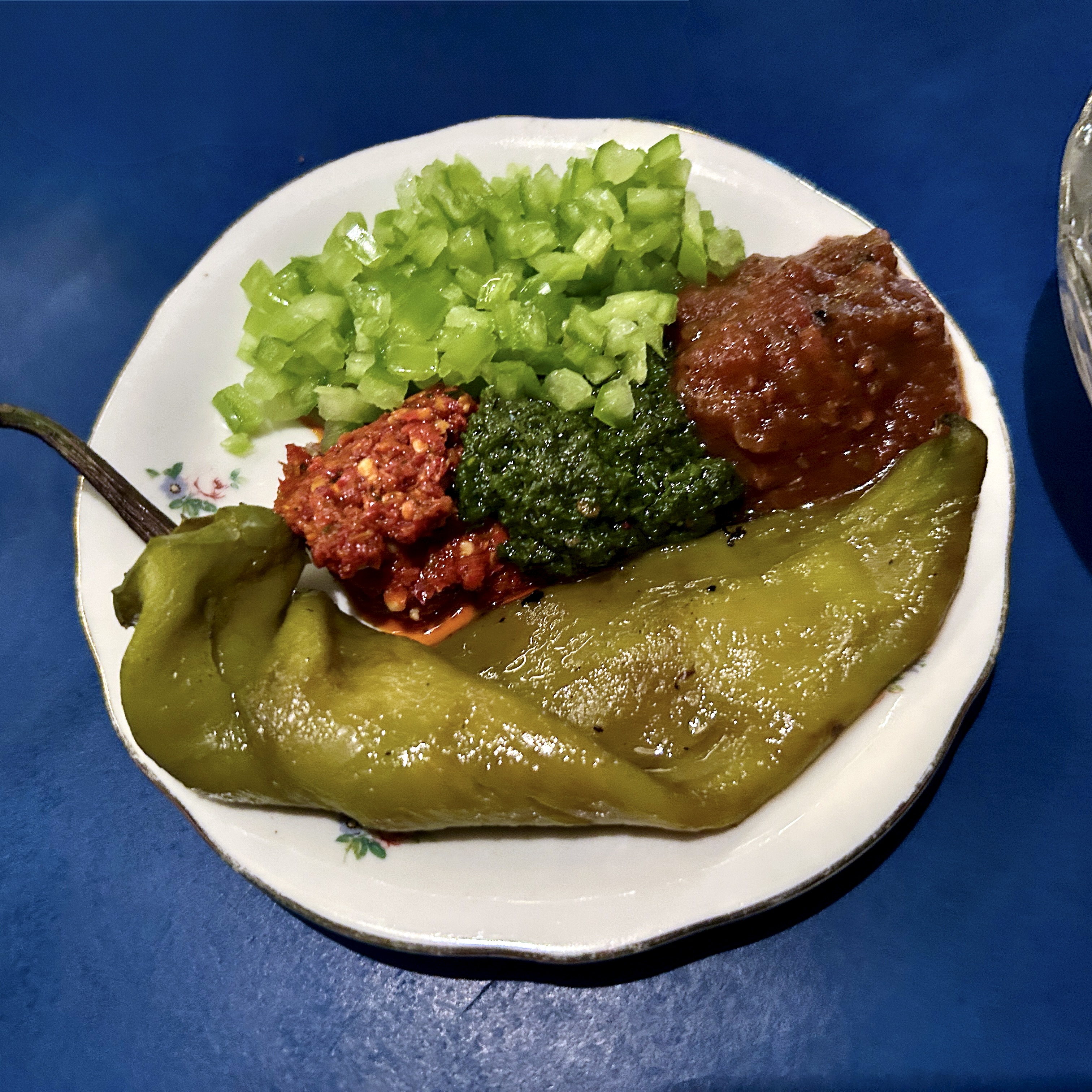 A small white floral plate with a whole roasted green pepper, chopped green vegetables, and three condiments — a red chili paste, a green herb sauce, and a brown sauce.