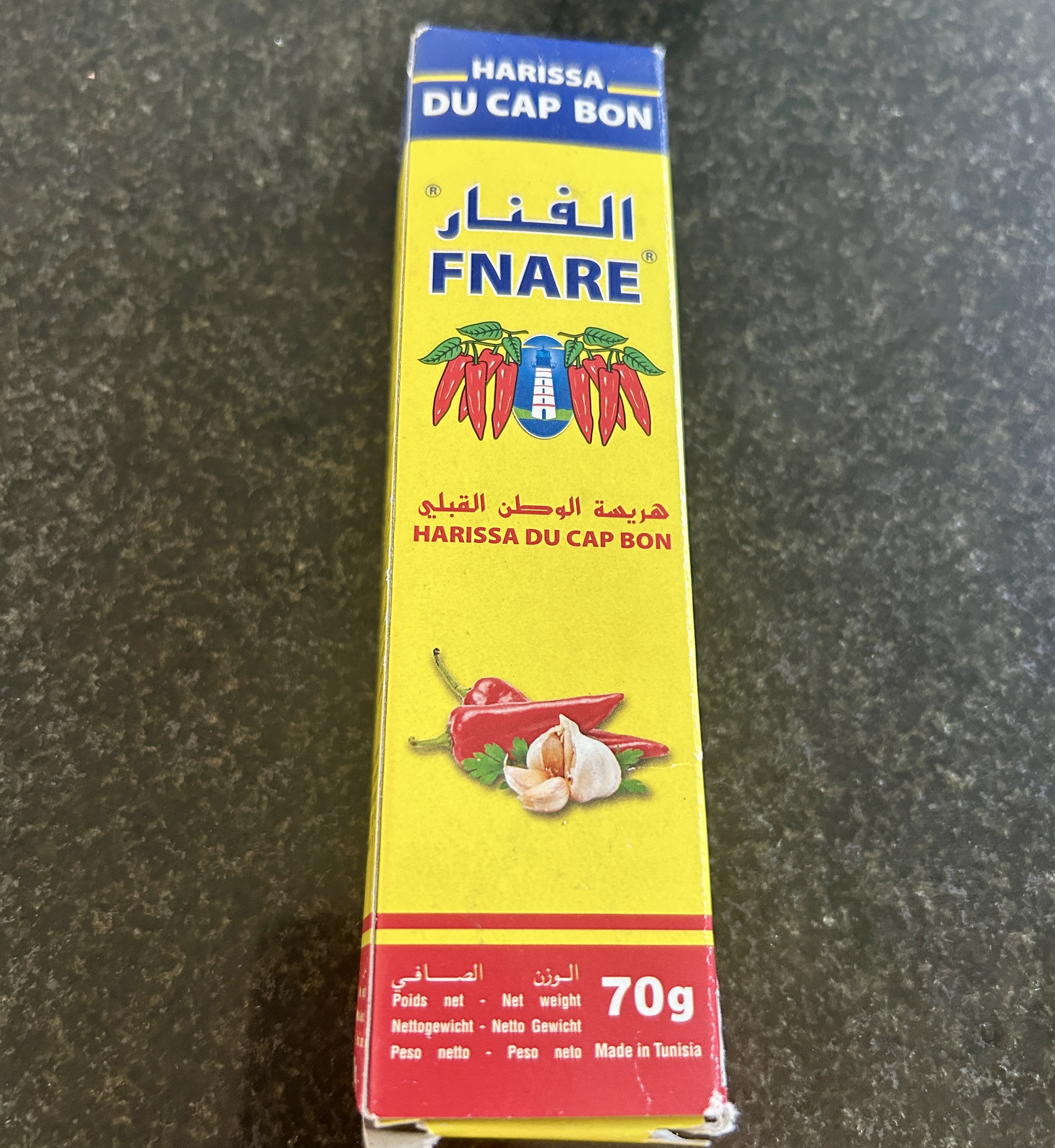 A yellow tube of Fnare Harissa du Cap Bon, made in Tunisia, with Arabic and French text and an illustration of red chili peppers and garlic.