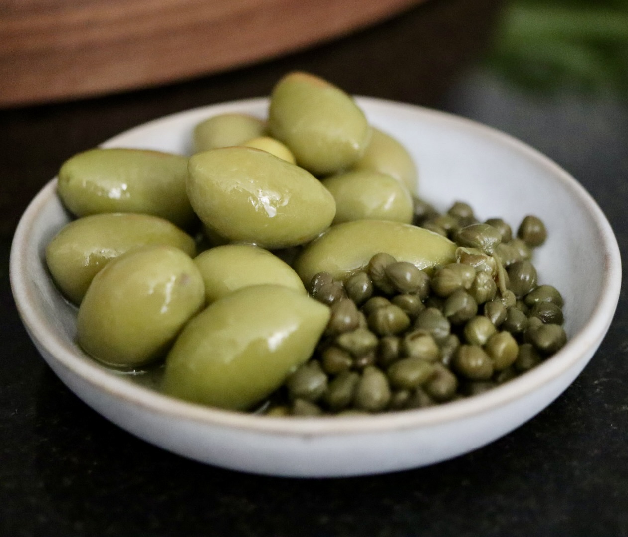 A white bowl with green olives and capers on a dark surface.