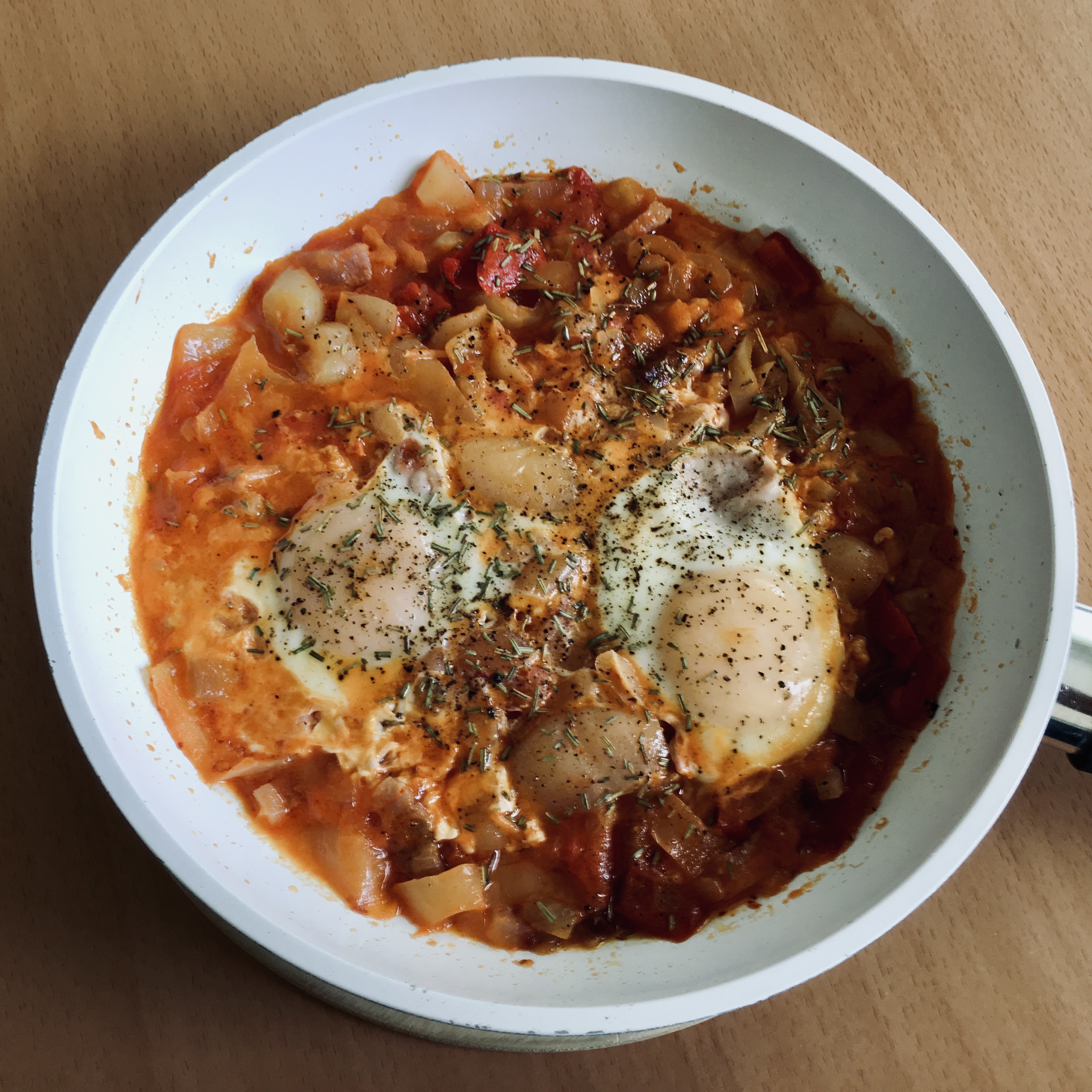 A white pan with lecho — a thick tomato and pepper stew — with two poached eggs on top, sprinkled with dried herbs and black pepper.
