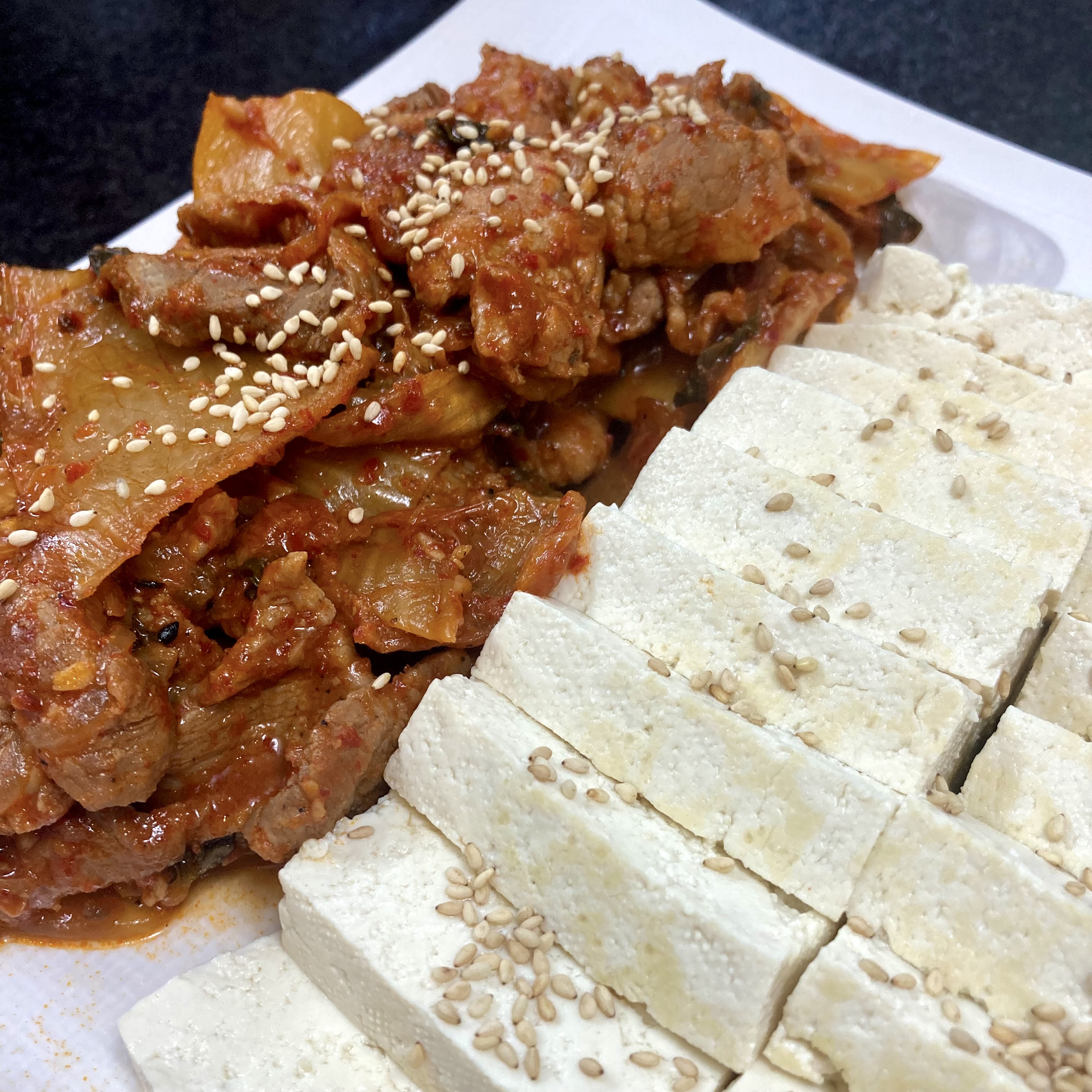 A white plate with sliced firm tofu sprinkled with sesame seeds alongside stir-fried kimchi and pork, also topped with sesame seeds.