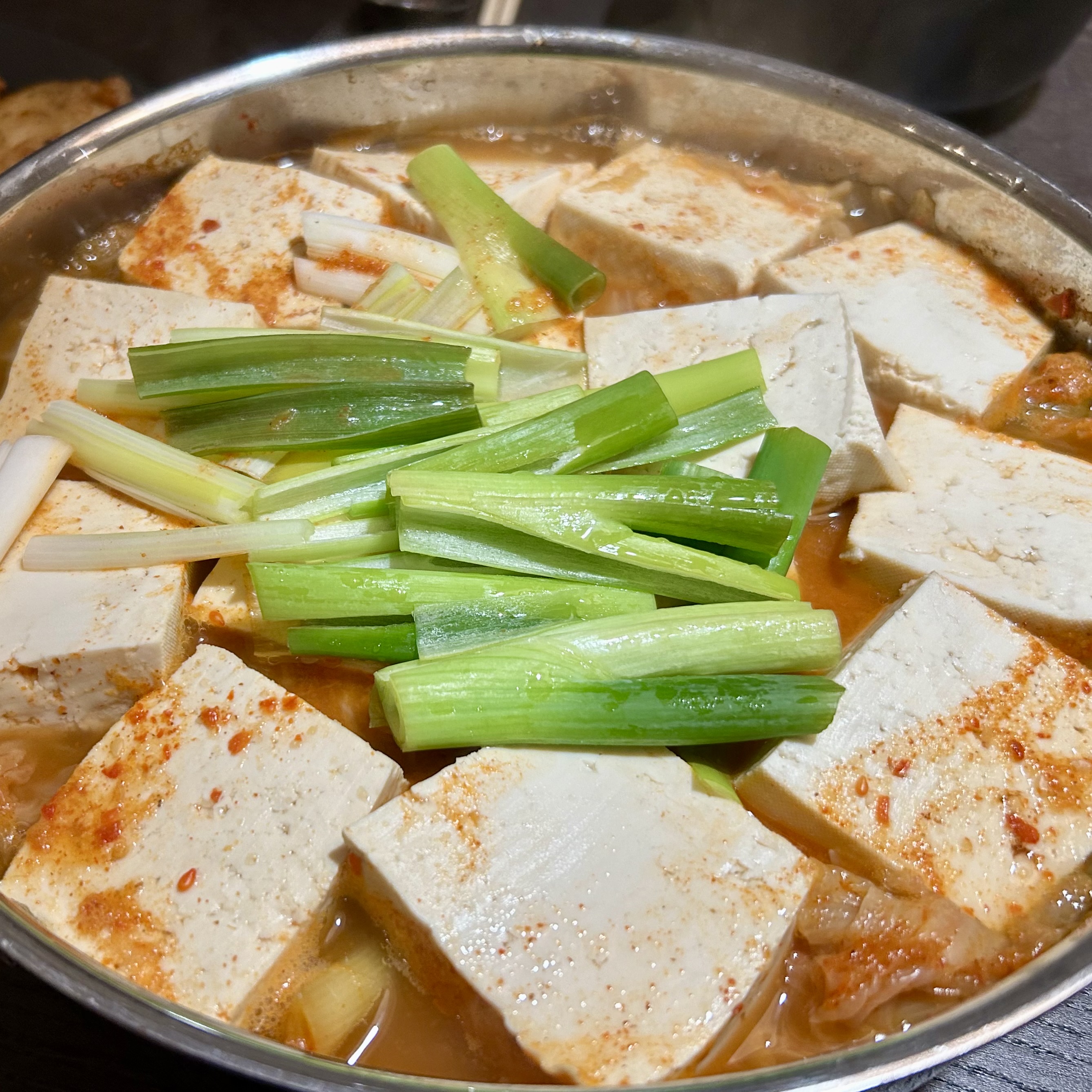 A large metal pan filled with tofu blocks in a reddish broth, topped with fresh green onion pieces, just before the final simmer.
