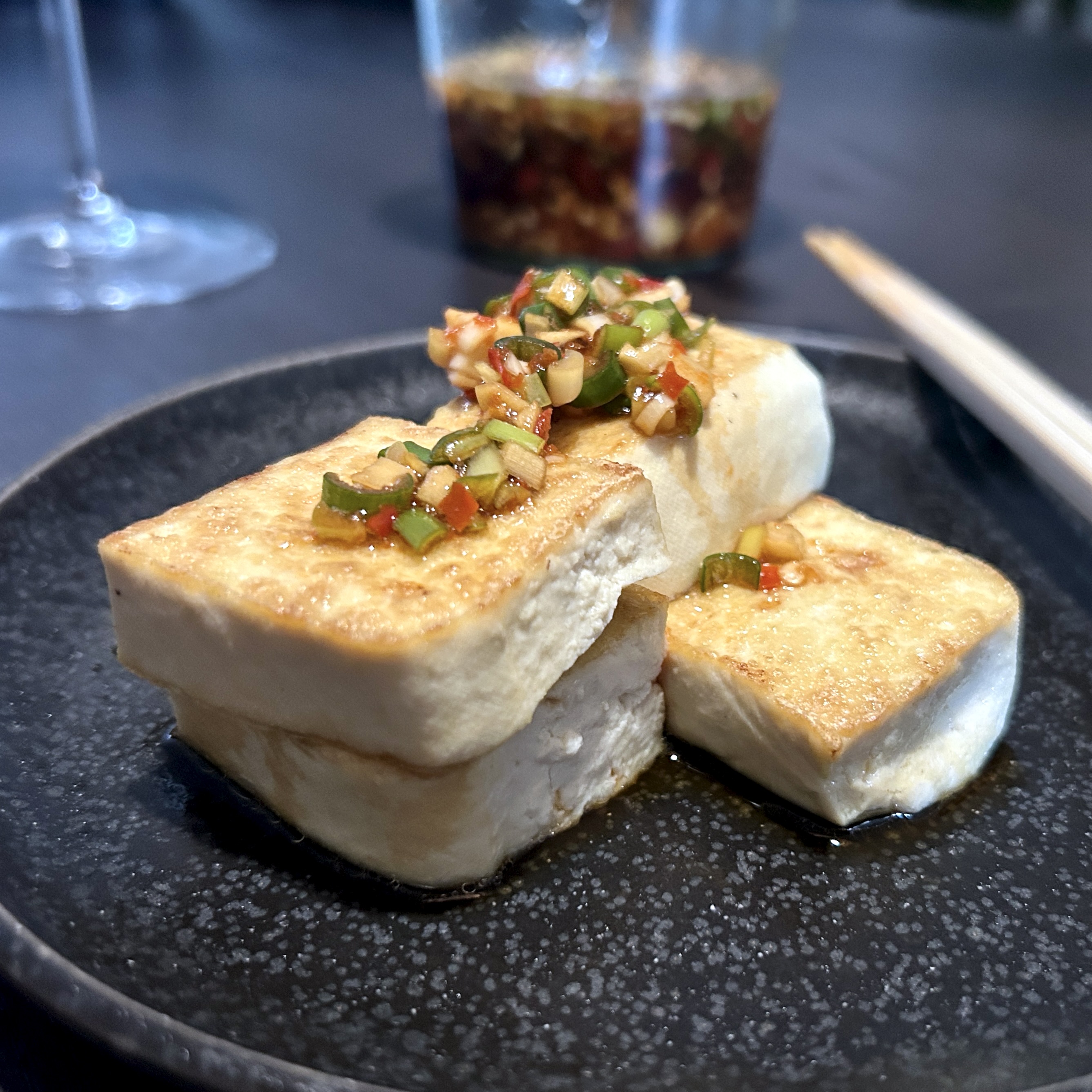 Pan-fried tofu blocks, golden on the outside, topped with a colorful yangnyeom ganjang of chopped chili, green onion, and garlic on a dark ceramic plate.