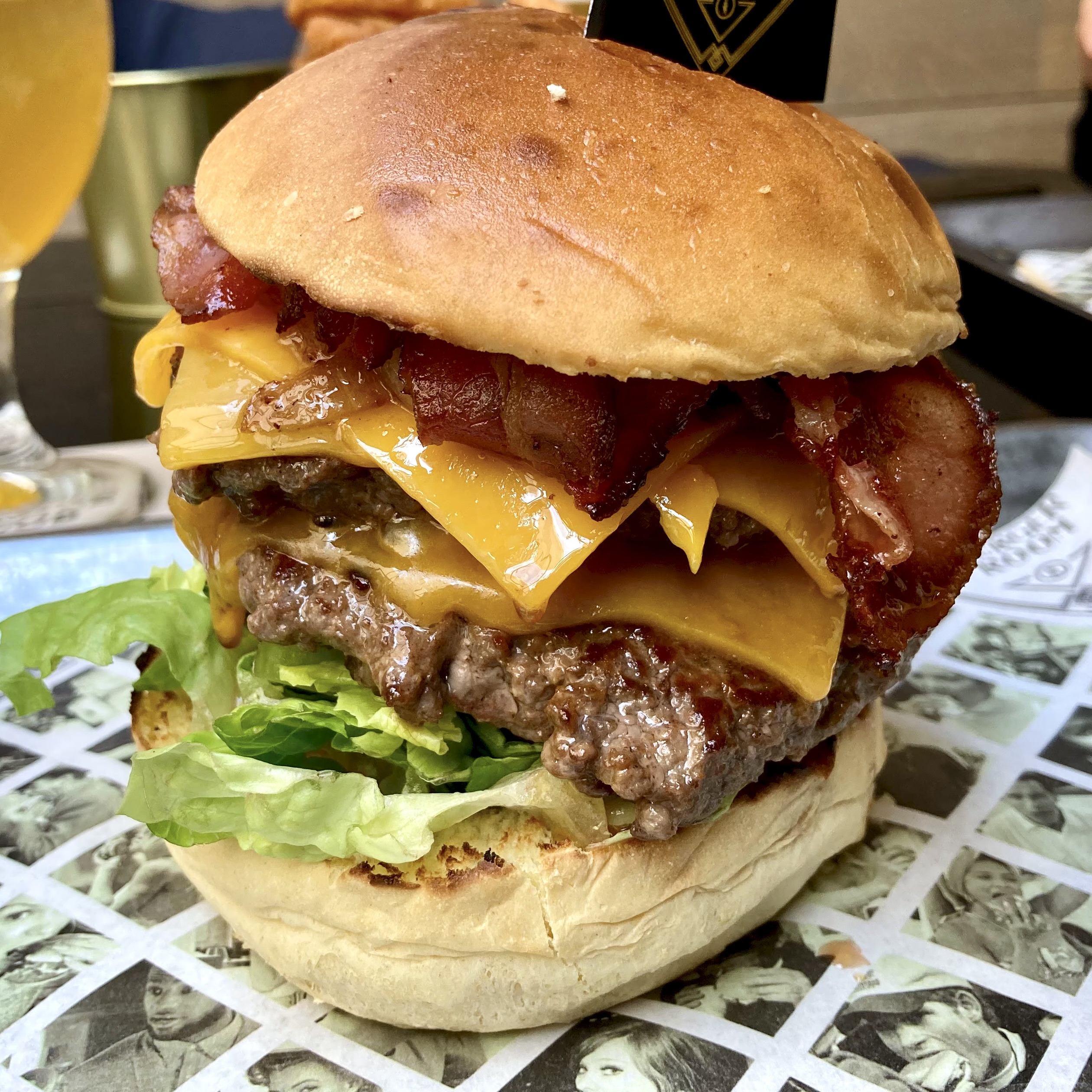 A large double cheeseburger with bacon, melted cheddar, and lettuce on a sesame bun, served on a printed paper wrapper.