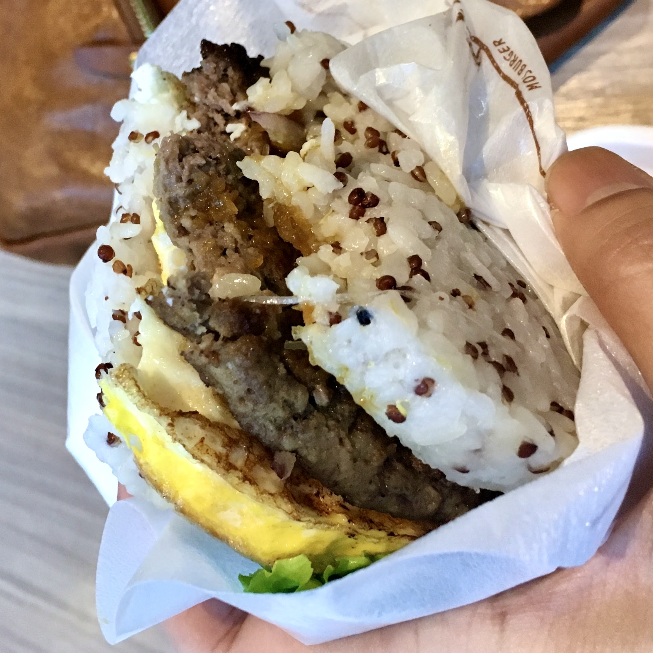 A hand holding a rice burger — a meat patty and fried egg sandwiched between two pressed rice buns, wrapped in paper.
