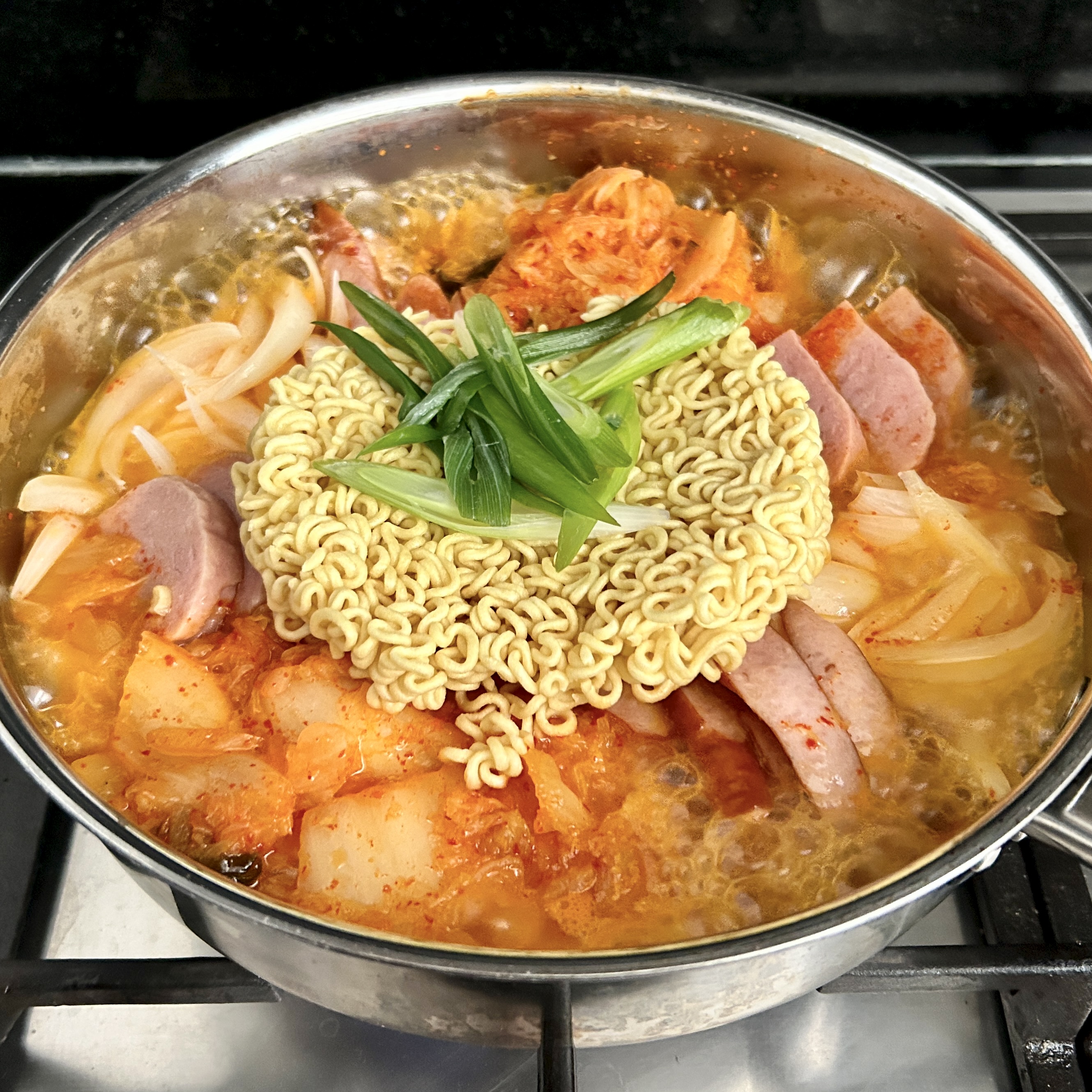 A large metal pan of bubbling budae-jjigae on a stovetop, with kimchi, spam, sliced sausage, udon noodles, ramyeon, and green onions.