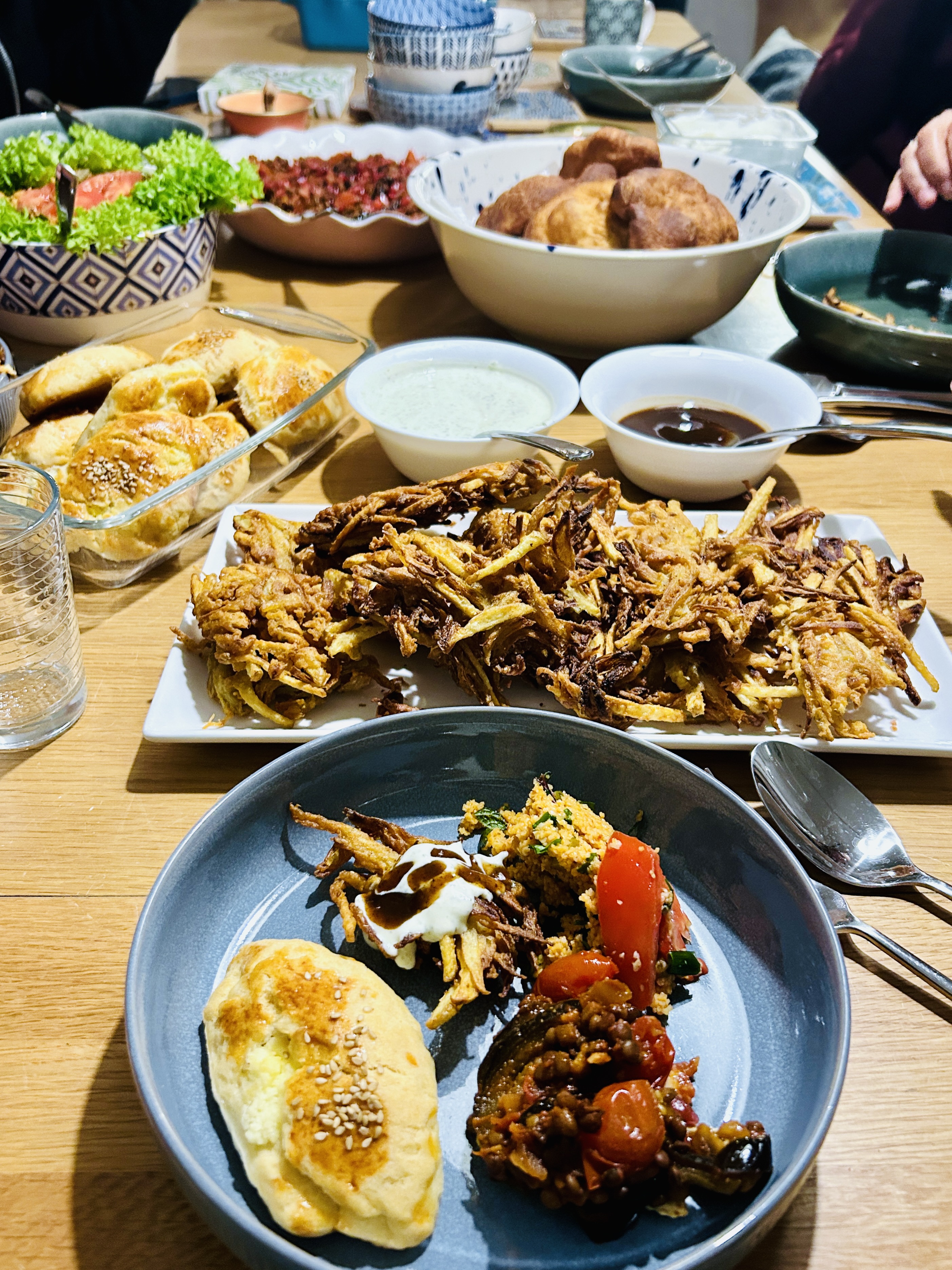 A shared dinner table with multiple dishes including fried onion fritters, pastries, two dipping sauces, a tomato salad, and a full plate in the foreground.