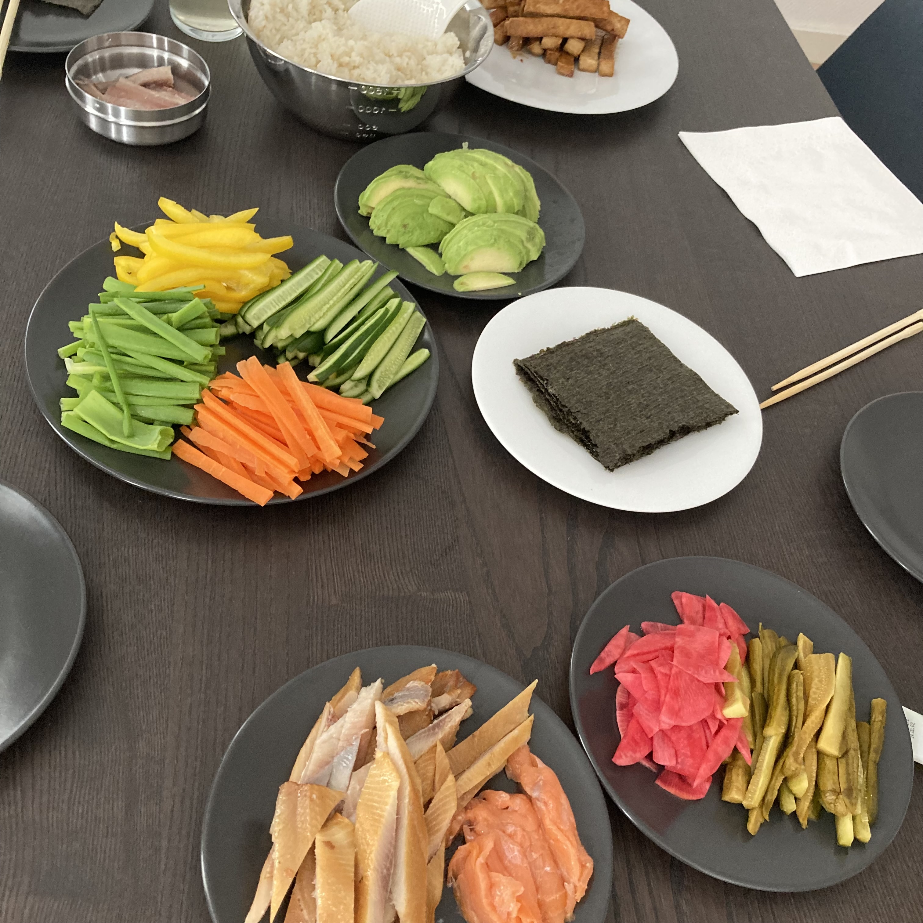 A table spread with gimbap and temaki ingredients: julienned vegetables, sliced avocado, smoked fish, pickled radish, nori sheets, and a bowl of rice.