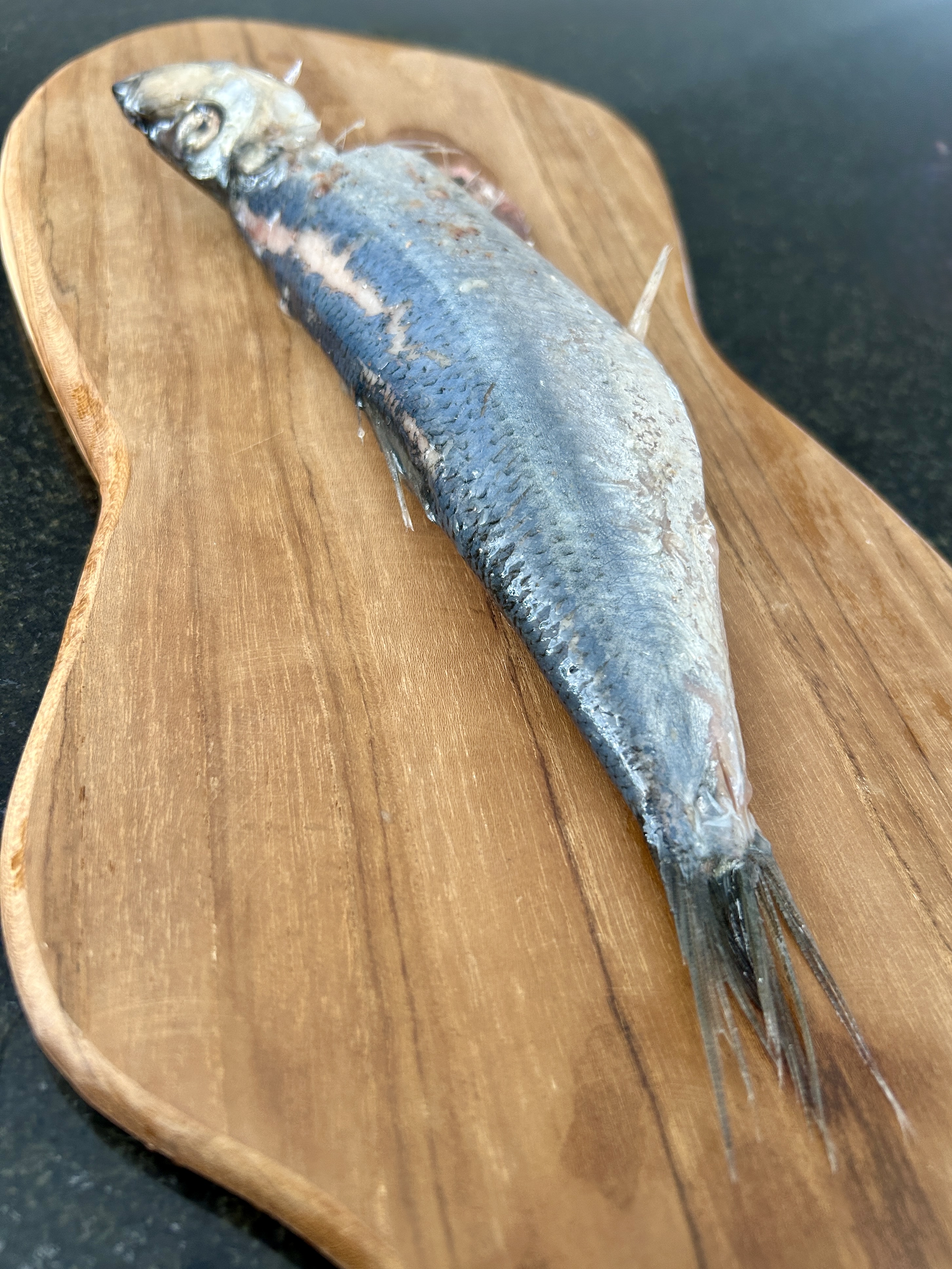 A whole uncleaned herring with head, scales, and tail intact, laid on a wooden cutting board.