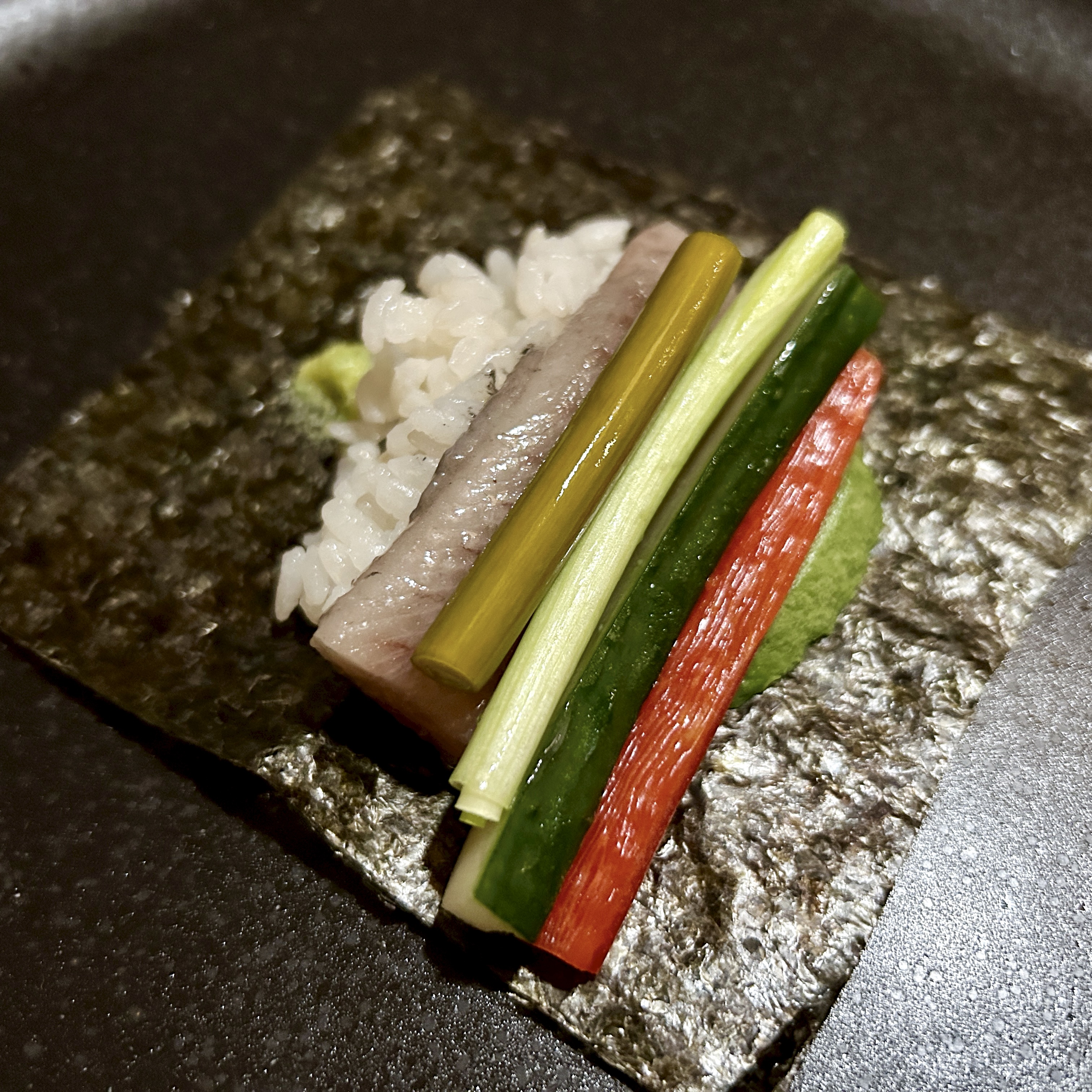 A sheet of nori laid flat with rice, a slice of herring, pickled vegetables, cucumber, and avocado arranged side by side, ready to be rolled.