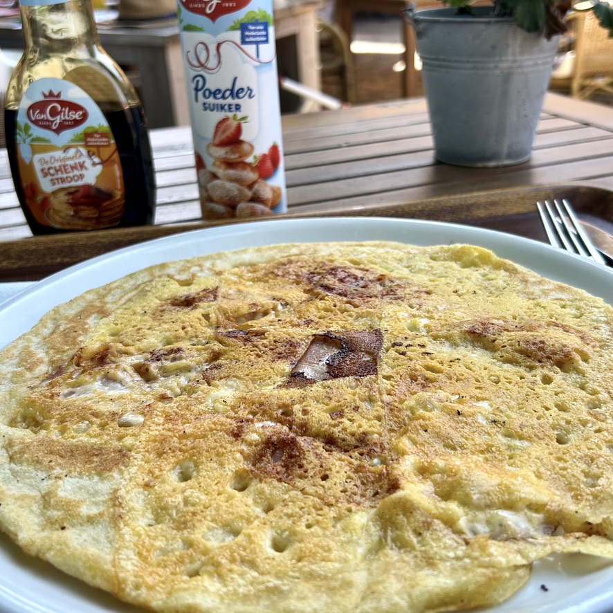 A large golden Dutch pannenkoek with bacon and cheese baked into the batter, served on a white plate with Dutch stroop and poedersuiker in the background.