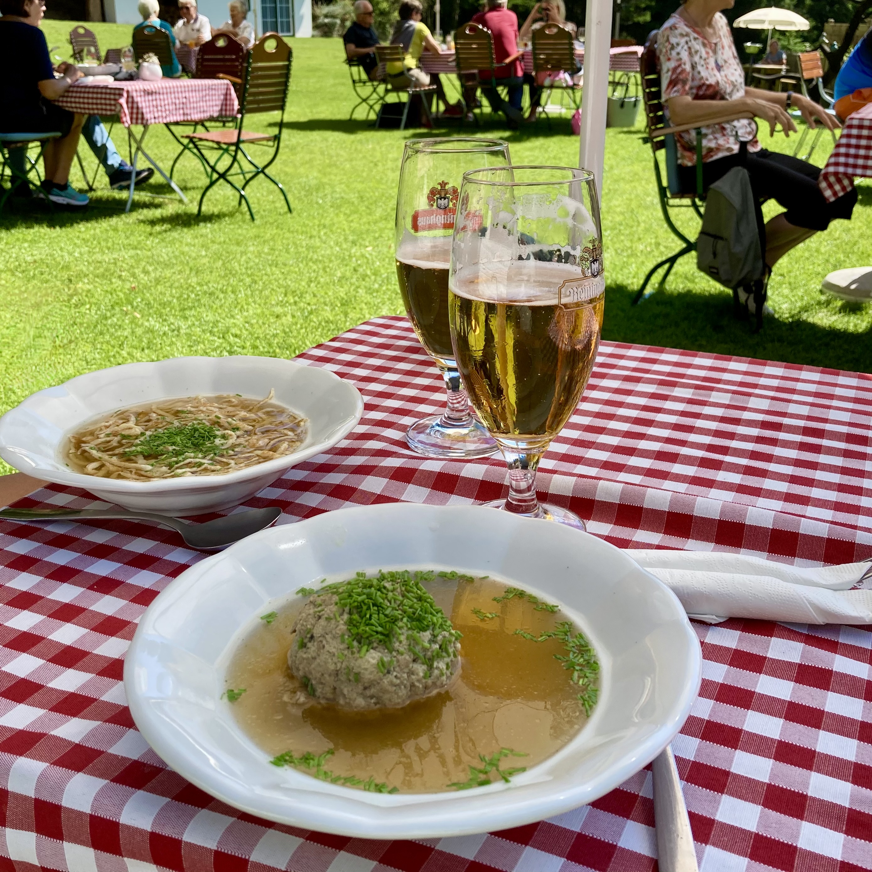 Two bowls of Austrian soup on a red-checked tablecloth at an outdoor garden restaurant — one with Frittatensuppe and one with a bread dumpling, with two glasses of beer alongside.
