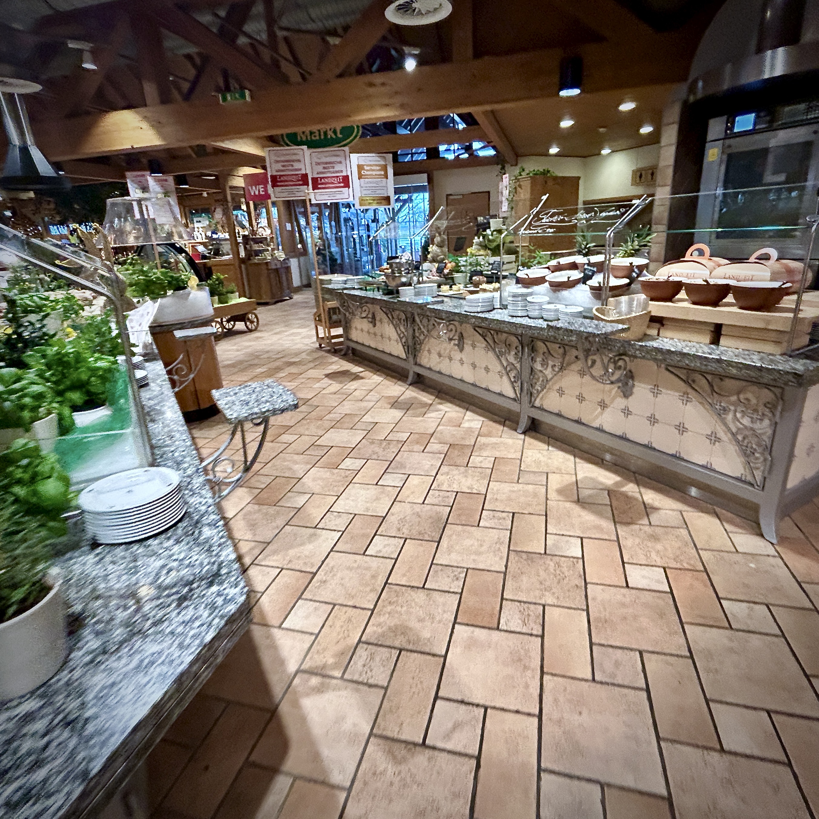 The interior of a Landzeit motorway rest stop in Austria, with a buffet counter, ornate ironwork, terra cotta tile floors, and a market area in the background.