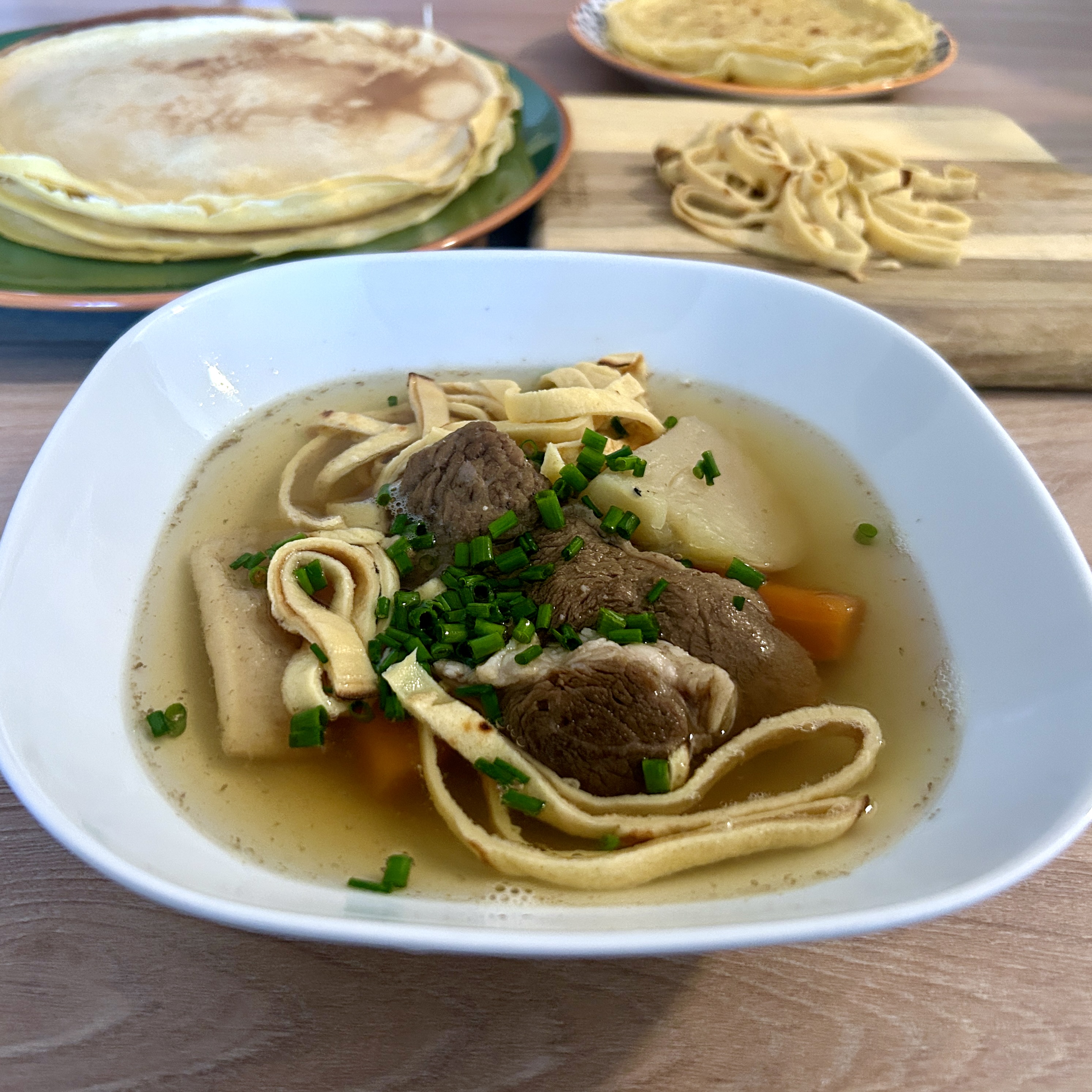 A white bowl of Hungarian húsleves with Fritatten pancake strips, beef, vegetables, and chopped chives, with a stack of palacsinta and sliced pancake strips visible in the background.