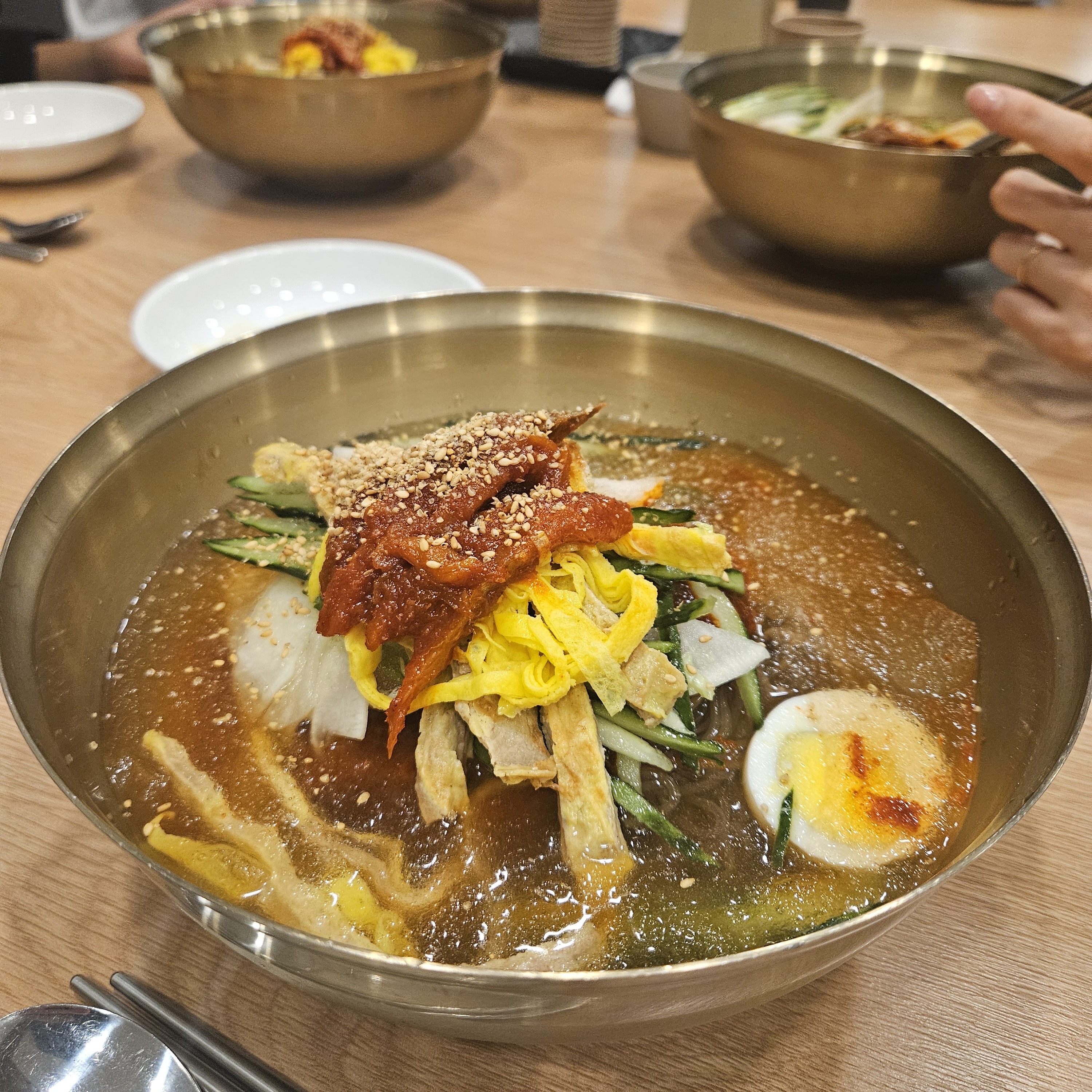 A large stainless steel bowl of naengmyeon — cold buckwheat noodles in broth topped with spicy beef, cucumber, egg, radish, and sesame seeds — with more bowls visible in the background.