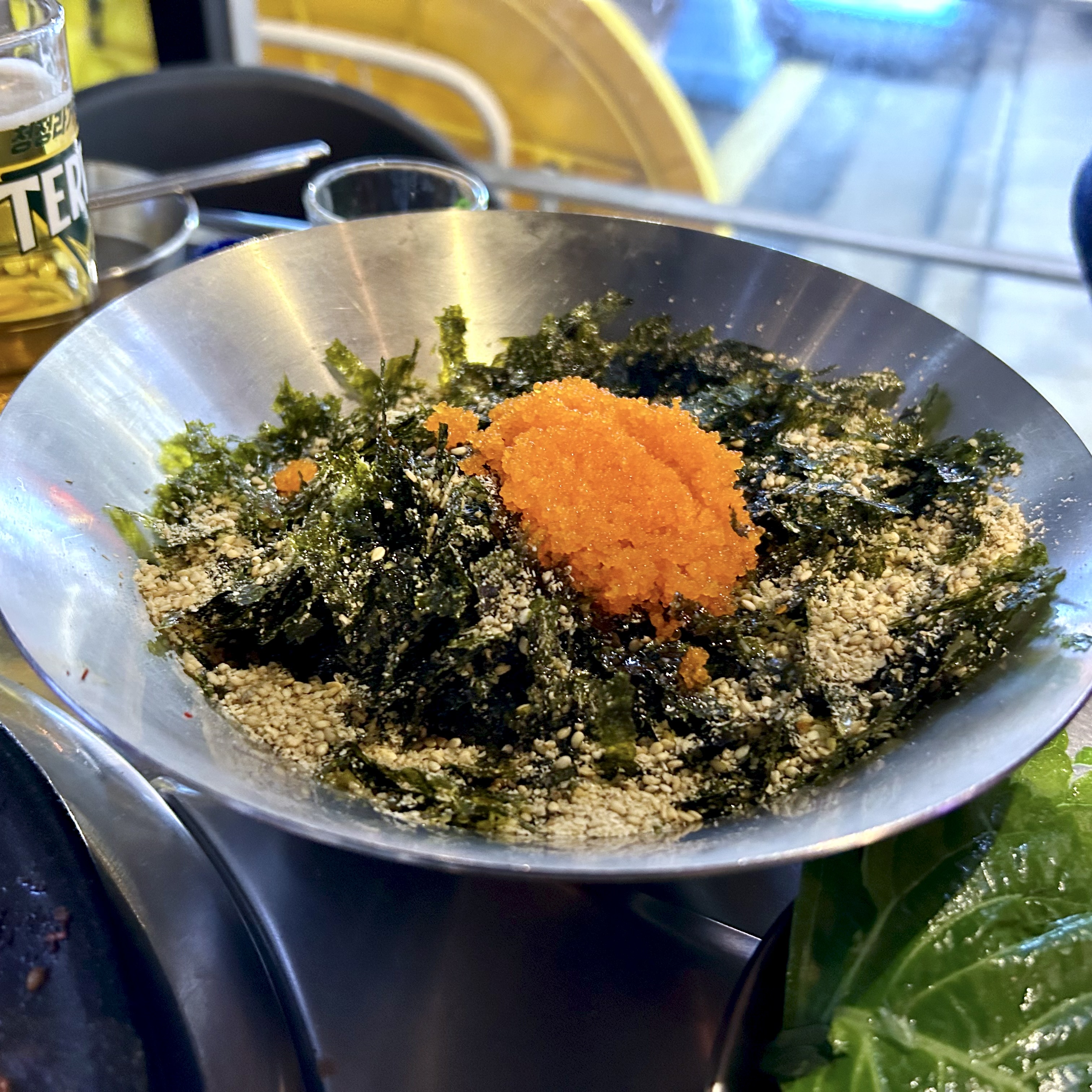 A stainless steel bowl with crumbled gim, sesame seeds, and a mound of bright orange fish roe, ready to be mixed by hand into rice.