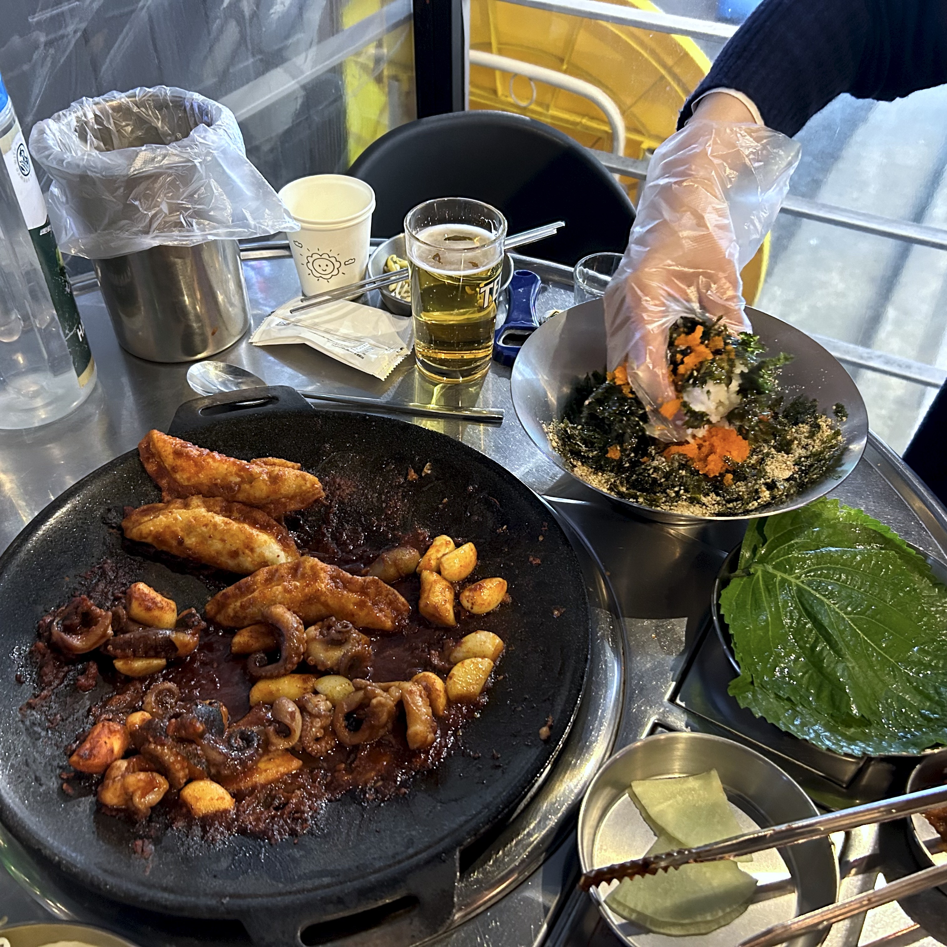 A table with a pan of jukkumi-bokkeum still on the burner, and a gloved hand mixing jumeokbap in a bowl alongside kkaennip leaves and a glass of beer.