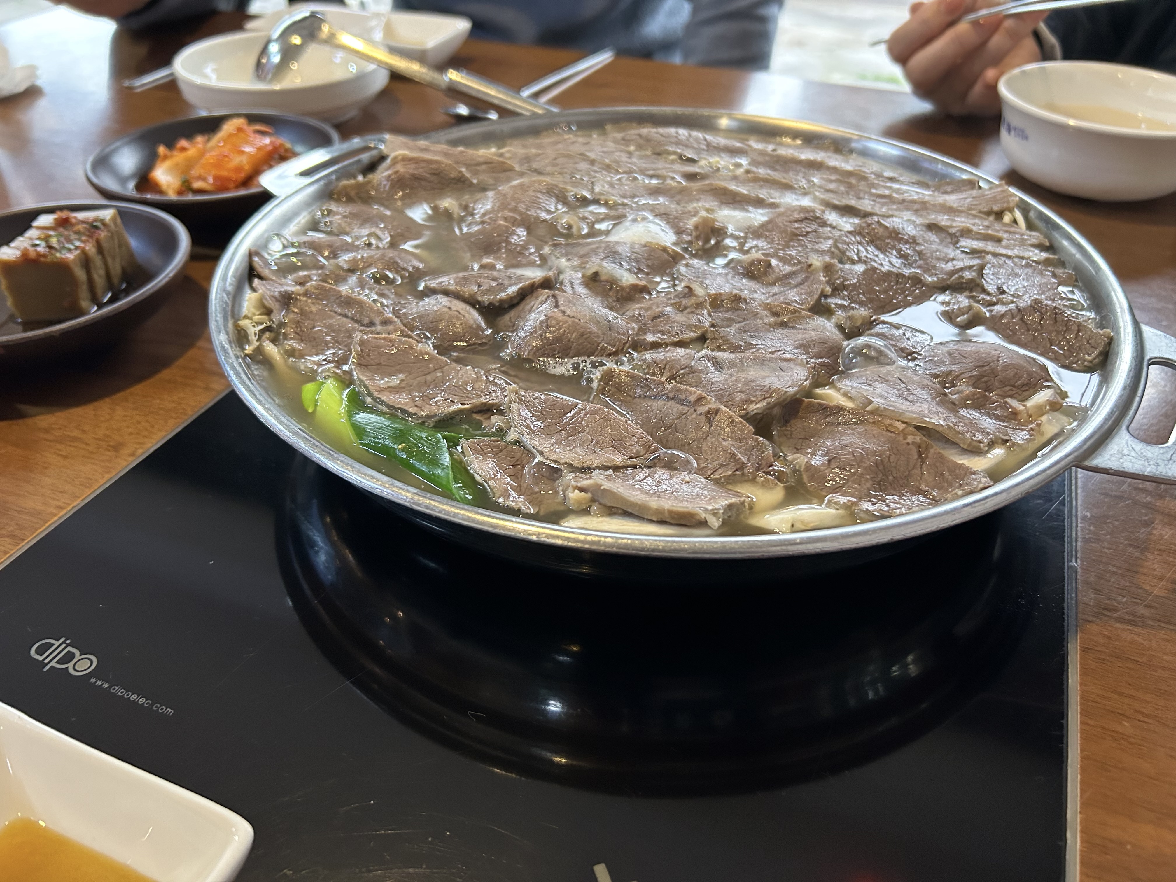 A large shallow pan filled with thin slices of beef simmering in clear broth on an induction burner, with green onion visible underneath.