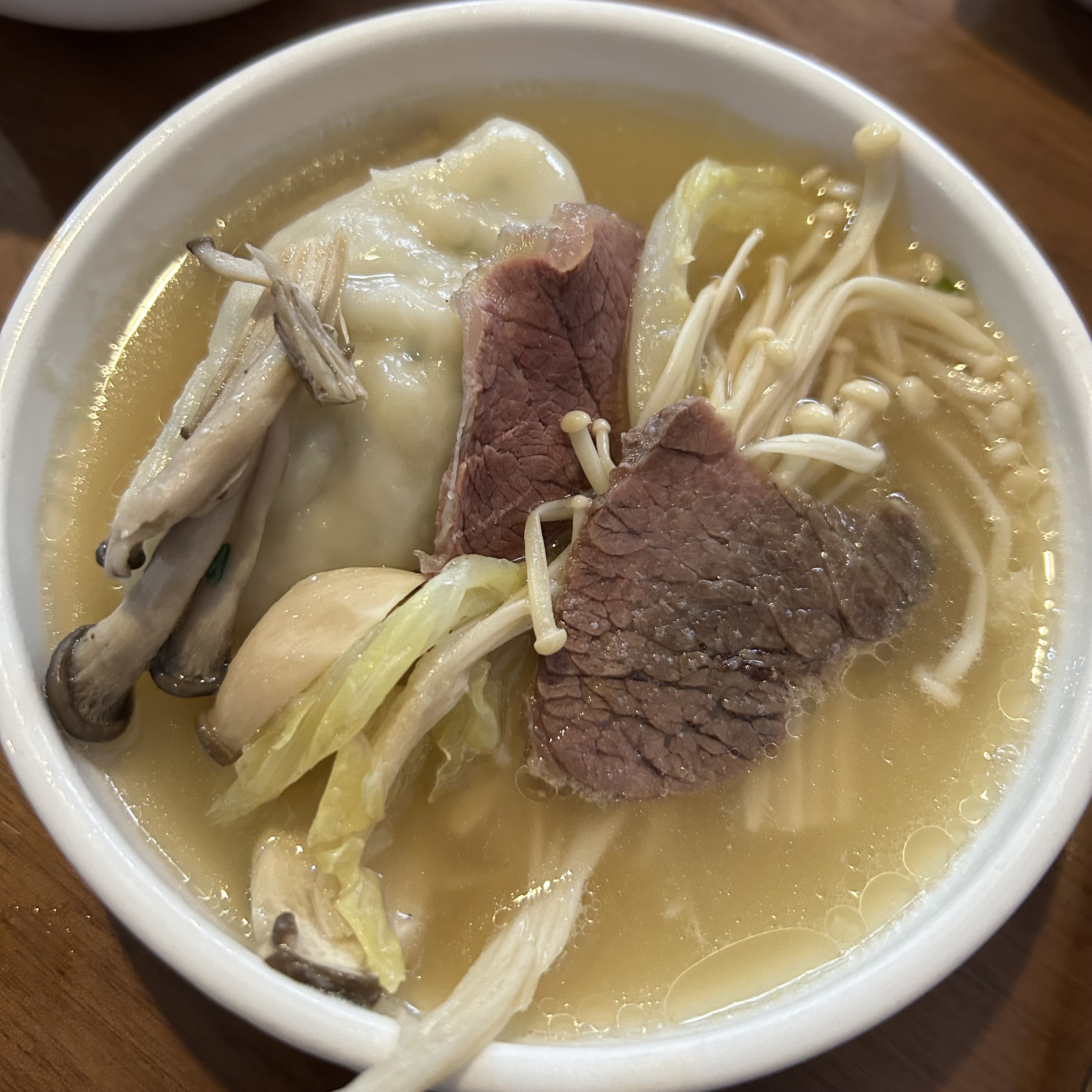 A white bowl of clear broth with mandu, sliced beef, mushrooms, enoki, and napa cabbage.